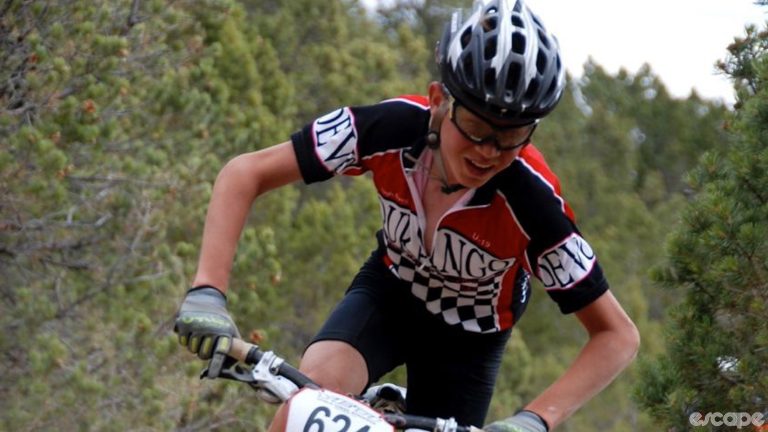 A young Sepp Kuss in Durango Devo kit races his mountain bike on a dusty trail with bushes in the background