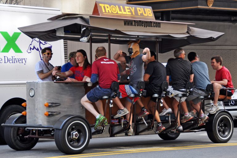 A beer bike full of rowdy men drives down a street. One of them is wearing a horse head mask.