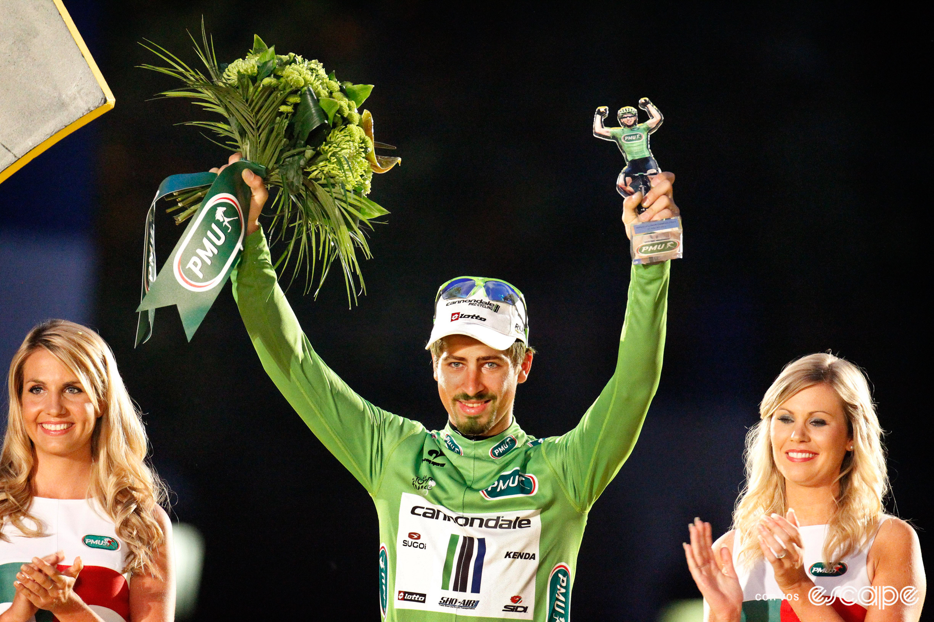 Peter Sagan on the final podium at the 2013 Tour de France, with two podium hostesses beside him, a bouquet of green flowers in one hand, and a trophy in the other.