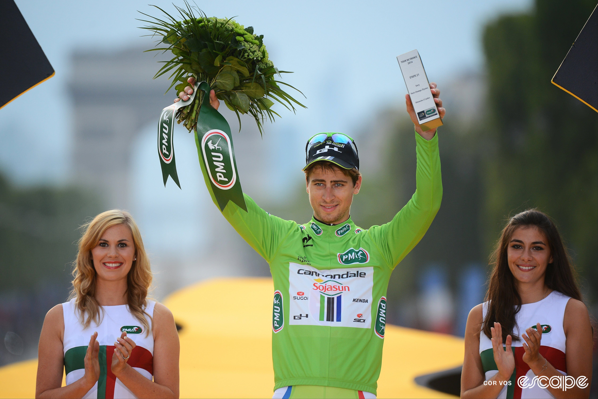 Peter Sagan on the final podium at the 2014 Tour de France wearing green, holding a bouquet of flowers and trophy, with a podium hostess on either side of him.