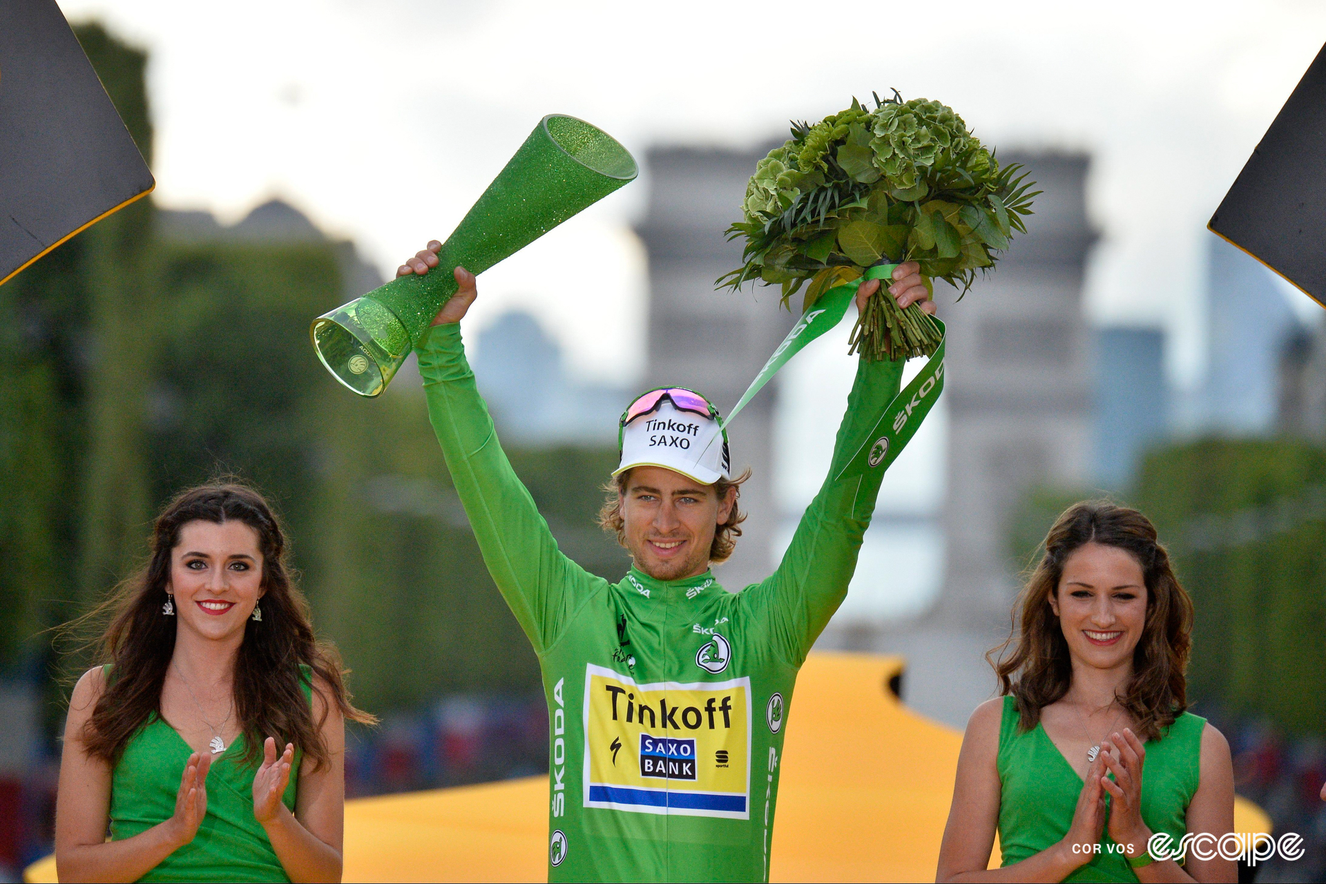 Peter Sagan on the final podium at the 2015 Tour de France wearing green, holding a bouquet of flowers and trophy, with a podium hostess on either side of him.