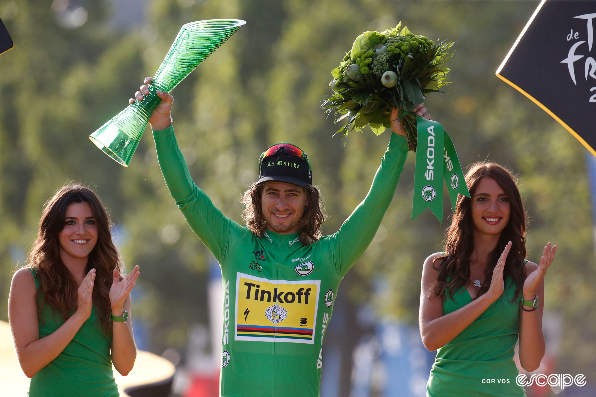 Peter Sagan on the final podium at the 2016 Tour de France wearing green, holding a bouquet of flowers and trophy, with a podium hostess on either side of him.