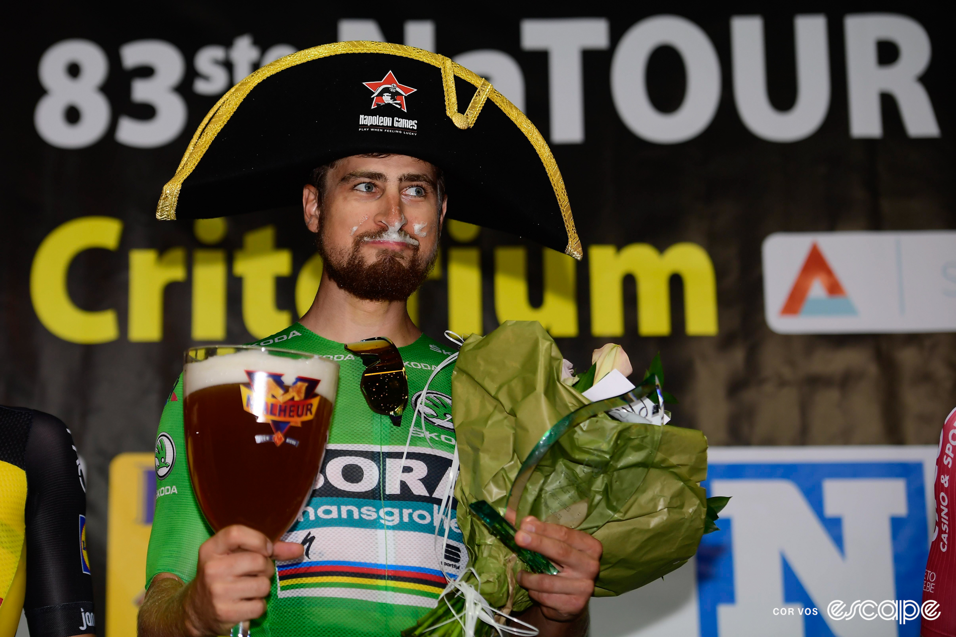 Peter Sagan stands on a podium with a Napoleon-esque hat on his head, beer froth on his face, and with a cheeky close-mouthed grin. He's holding a giant beer and a bouquet of flowers.