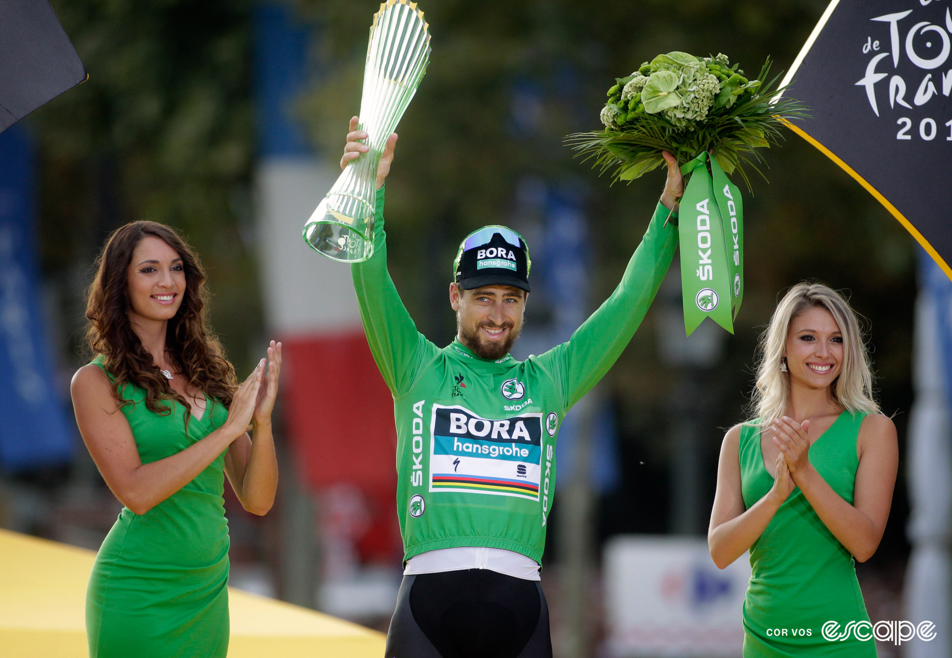 Peter Sagan on the final podium at the 2018 Tour de France wearing green, holding a bouquet of flowers and trophy, with a podium hostess on either side of him.