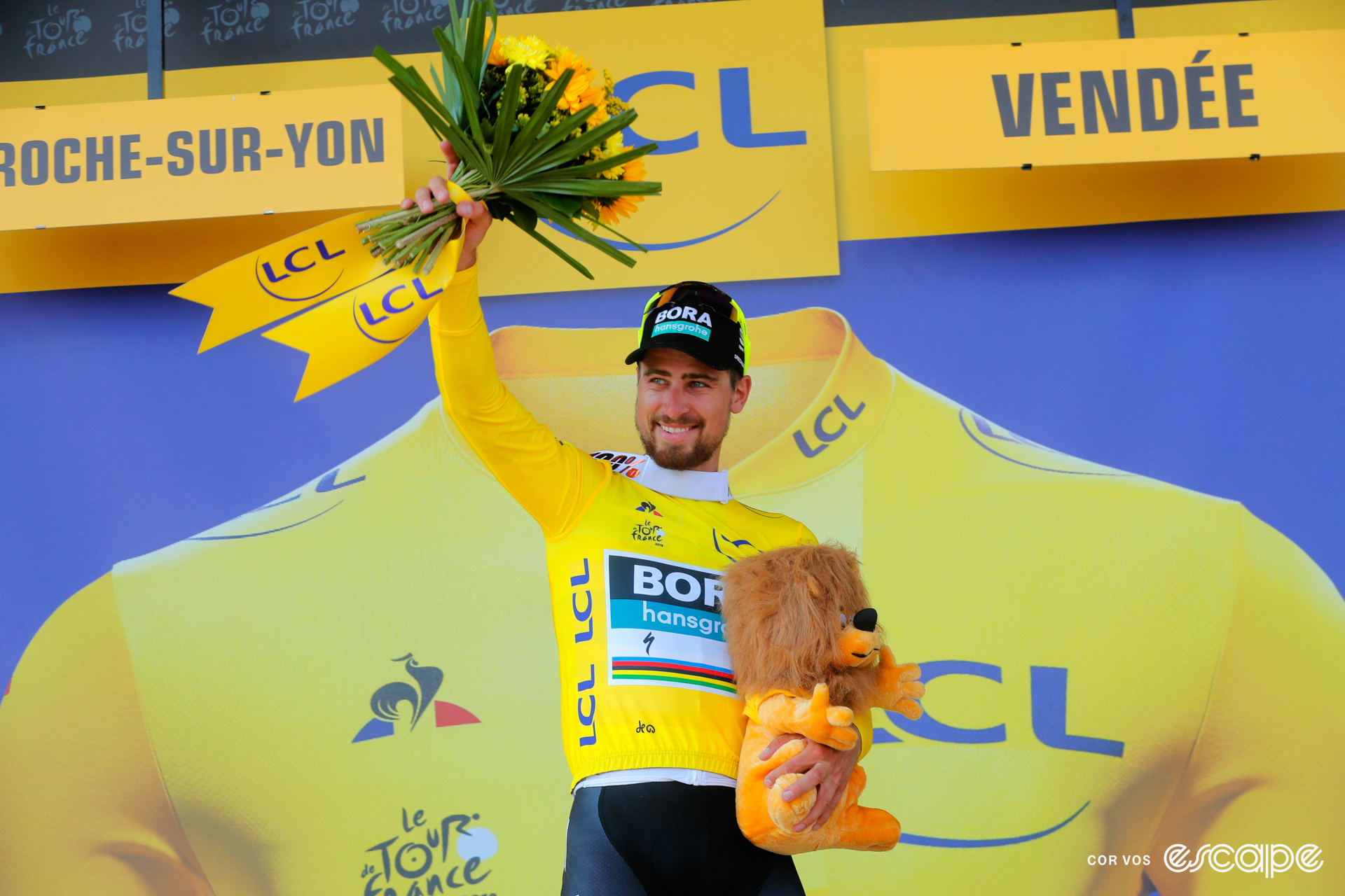 Peter Sagan stands on the podium after a Tour de France stage, wearing the leader's yellow jersey, holding a bouquet of flowers above his head, and holding a yellow line to his chest with his left hand.