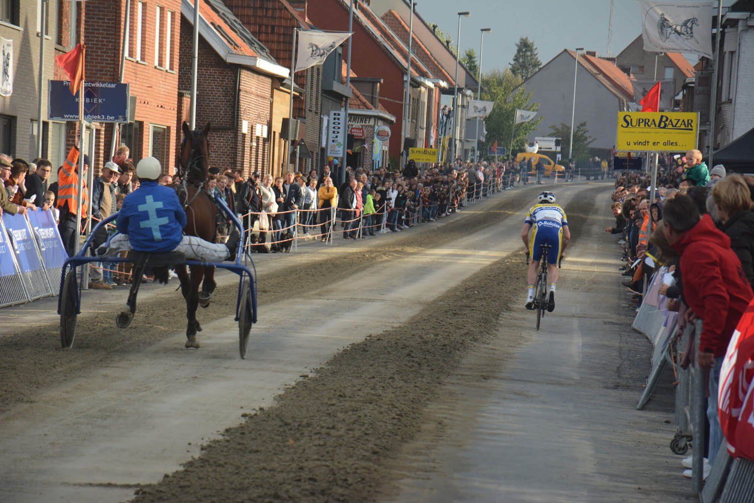 A cyclist rides up one side of a sandy street, while a horse and buggy race up the other. 