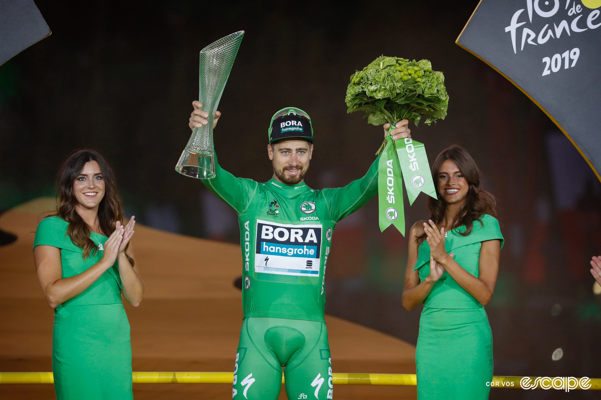 Peter Sagan on the final podium at the 2019 Tour de France wearing green, holding a bouquet of flowers and trophy, with a podium hostess on either side of him.