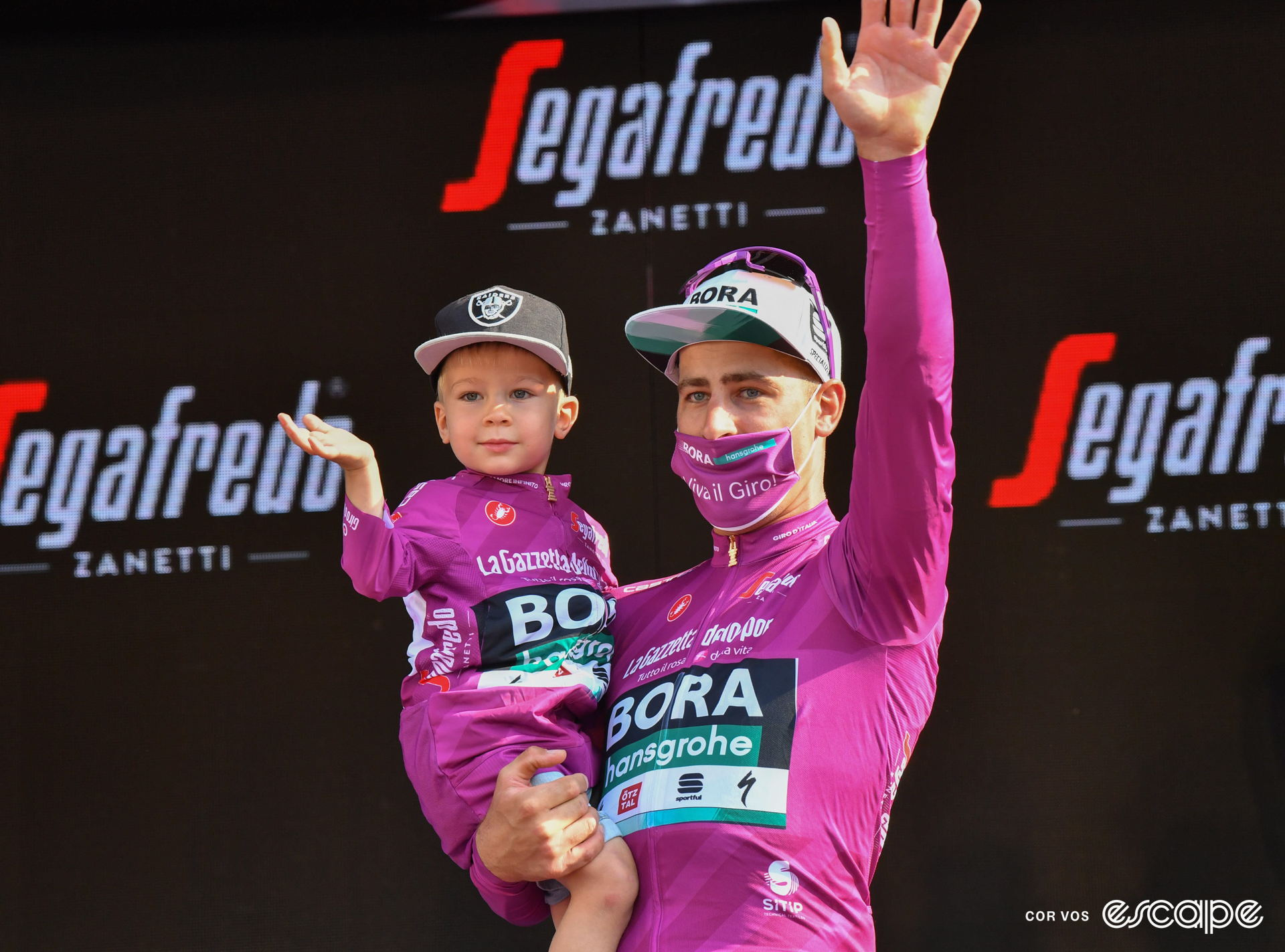 Peter Sagan stands on the final podium at the 2021 Giro d'Italia, dressed in the purple jersey of points classification leader, wearing a purple mask on his face, and holding his young son who's also in purple.