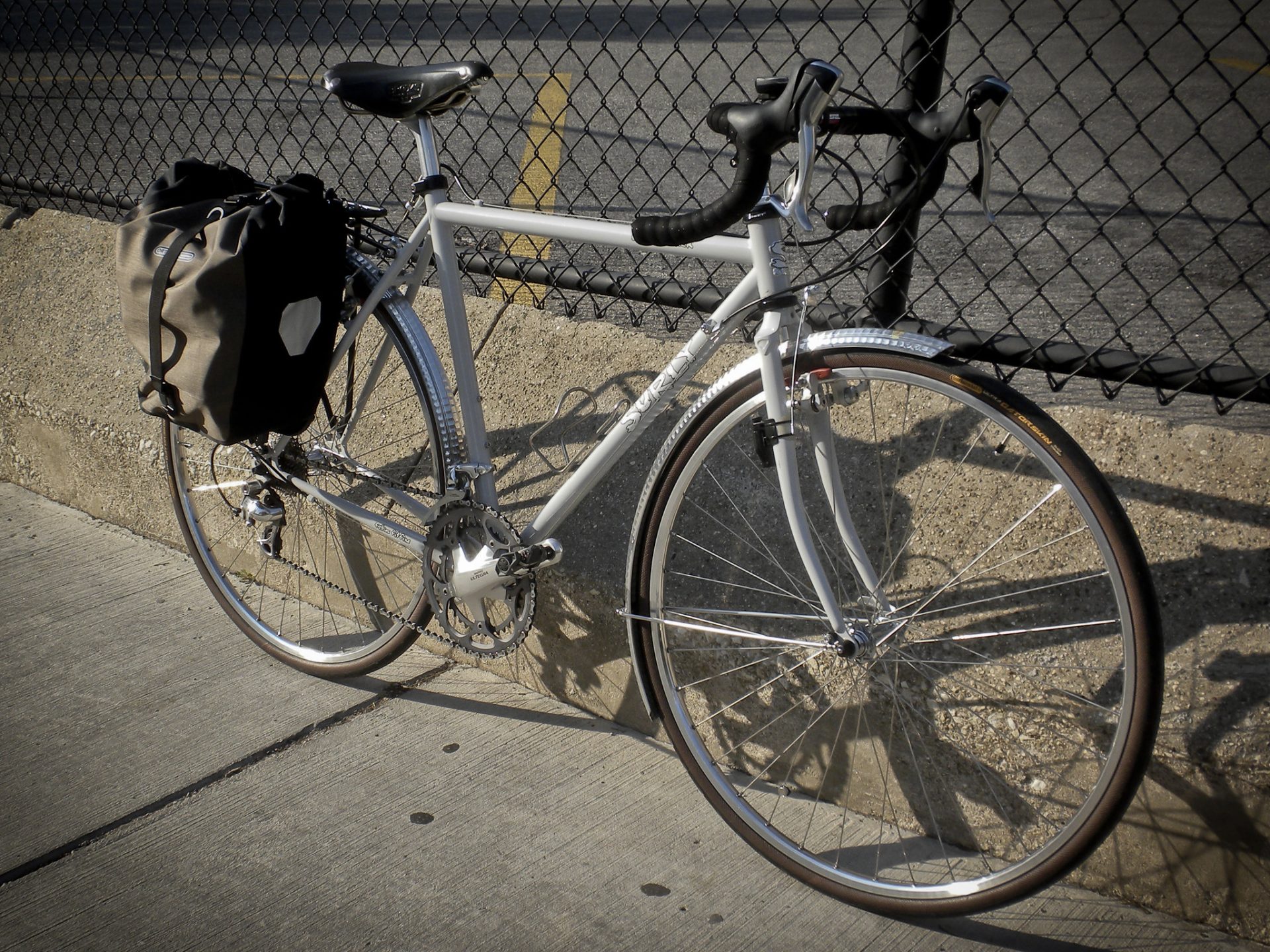 A white Surly Cross-Check leaning against a fence. It is built up with road gearing, a drop handlebar, full length hammered metal mudguards, a pannier rack and a Brooks saddle. 