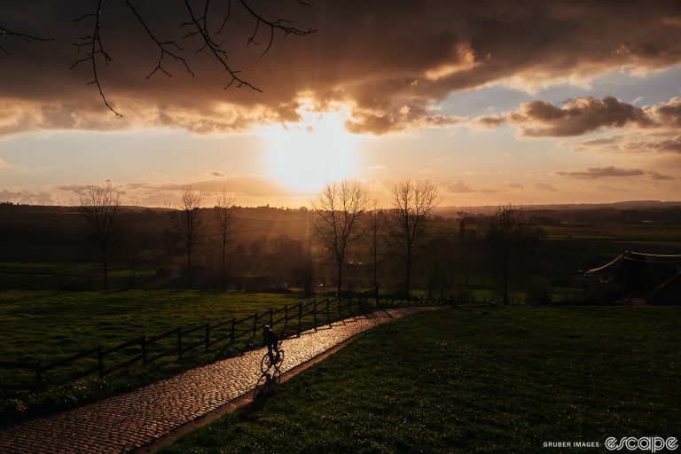 A lone rider climbs a cobbled road. A low sun behind the rider shines a golden light across the cobbles, but the rider and the background - trees without leaves and a Belgian farmscape - are shown in silhouette.