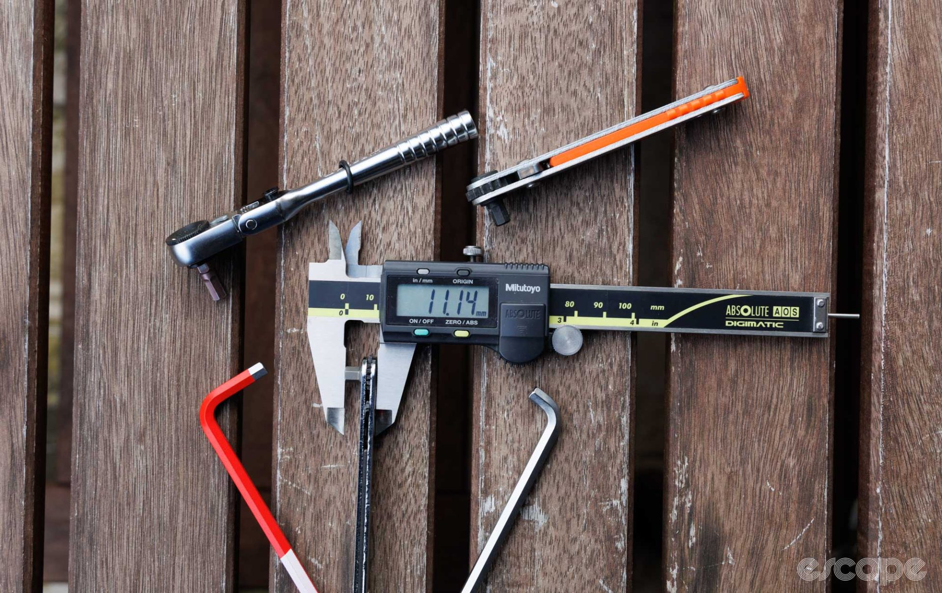 A handful of ratchet and non-ratchet hex tools on a bench. In the centre sits a low profile ratchet being measured by digital calipers. The calipers read 11.14 mm. 