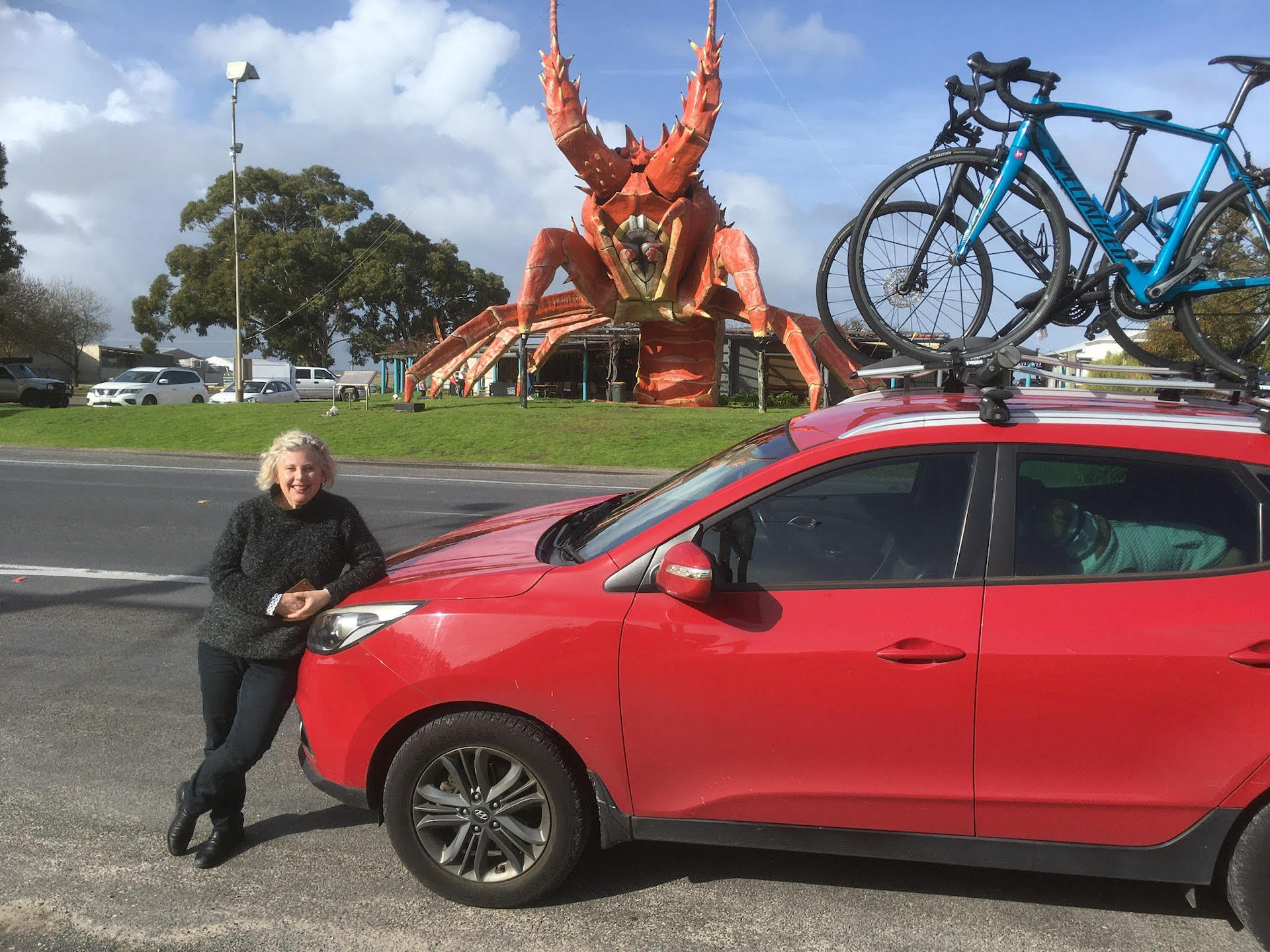 A woman leans against a car which has two bikes mounted on the roof. In the background is the Big Lobster, a giant statue of a red lobster.