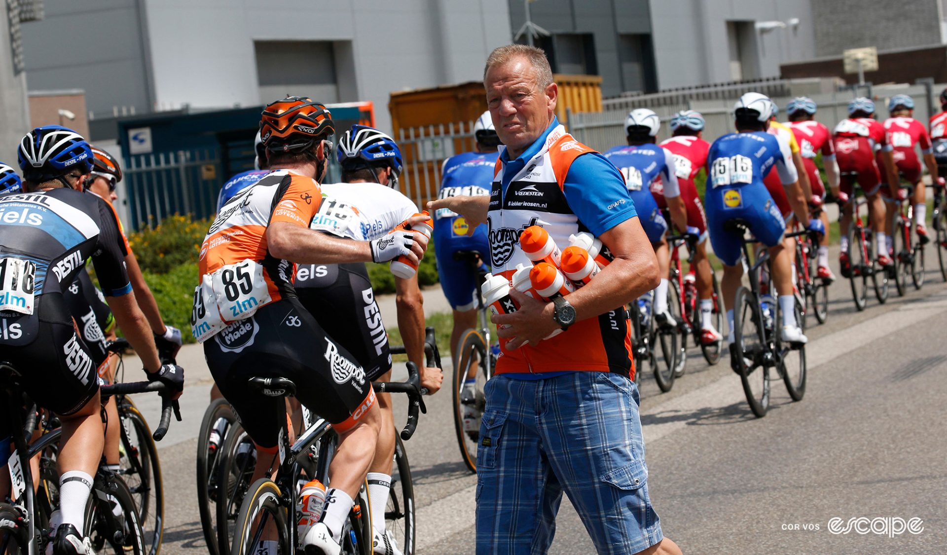 Soigneur Geert van Diepen hands bottles to riders at a race. The Belgian soigneur is holding six bottles in his left hand and crook of his arm as he hands one off in a no-look transfer that would make Aaron Rodgers weep.