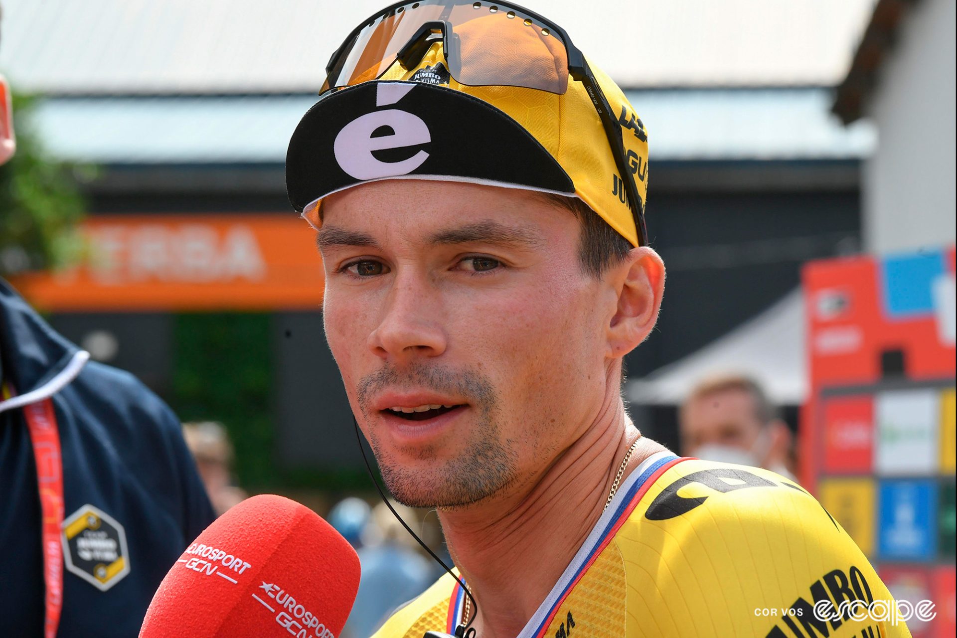 Primož Roglič in profile, speaking to a TV microphone at a race. He's wearing a Jumbo team cap with a large Cervelo E.
