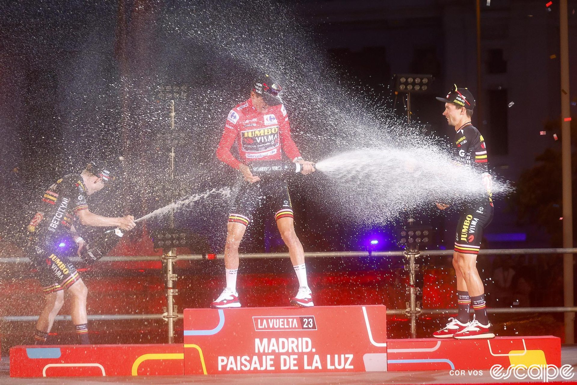 Sepp Kuss sprays champagne at teammates Primož Roglič (right) and Jonas Vingegaard (left) on the podium for the 2023 Vuelta a España. All three riders have champagne bottles and are dousing each other, which Richard Plugge approves of as long as they don't drink it.