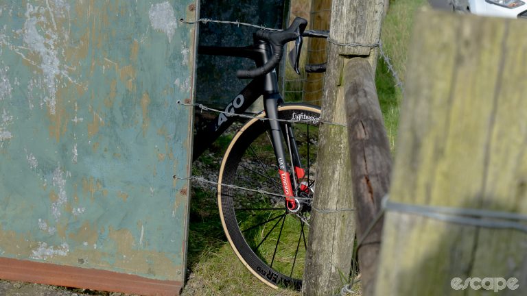 The photo shows the front half of a Colnago V4Rs with Lightweight wheels perched against the side of an old bus stop shelter and a barb wire fence.