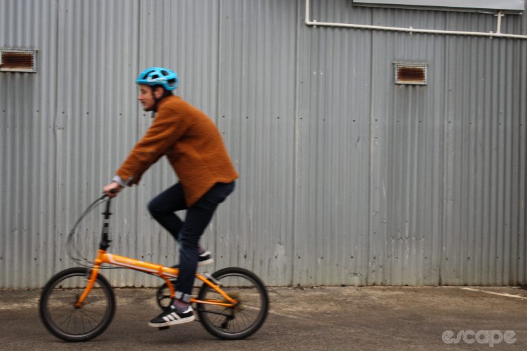The author rides his basic orange folding bike past a metal wall, motion-blurred.