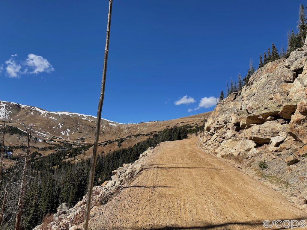 Old Fall River Road rises away from the camera. The dirt road is steeply pitched, with a 20-foot tall grey wooden pole on the left side as a marker for plows in spring. Ahead, the Trail Ridge visitor center sits perched high above on a ridgeline crested with a snowy cornice.