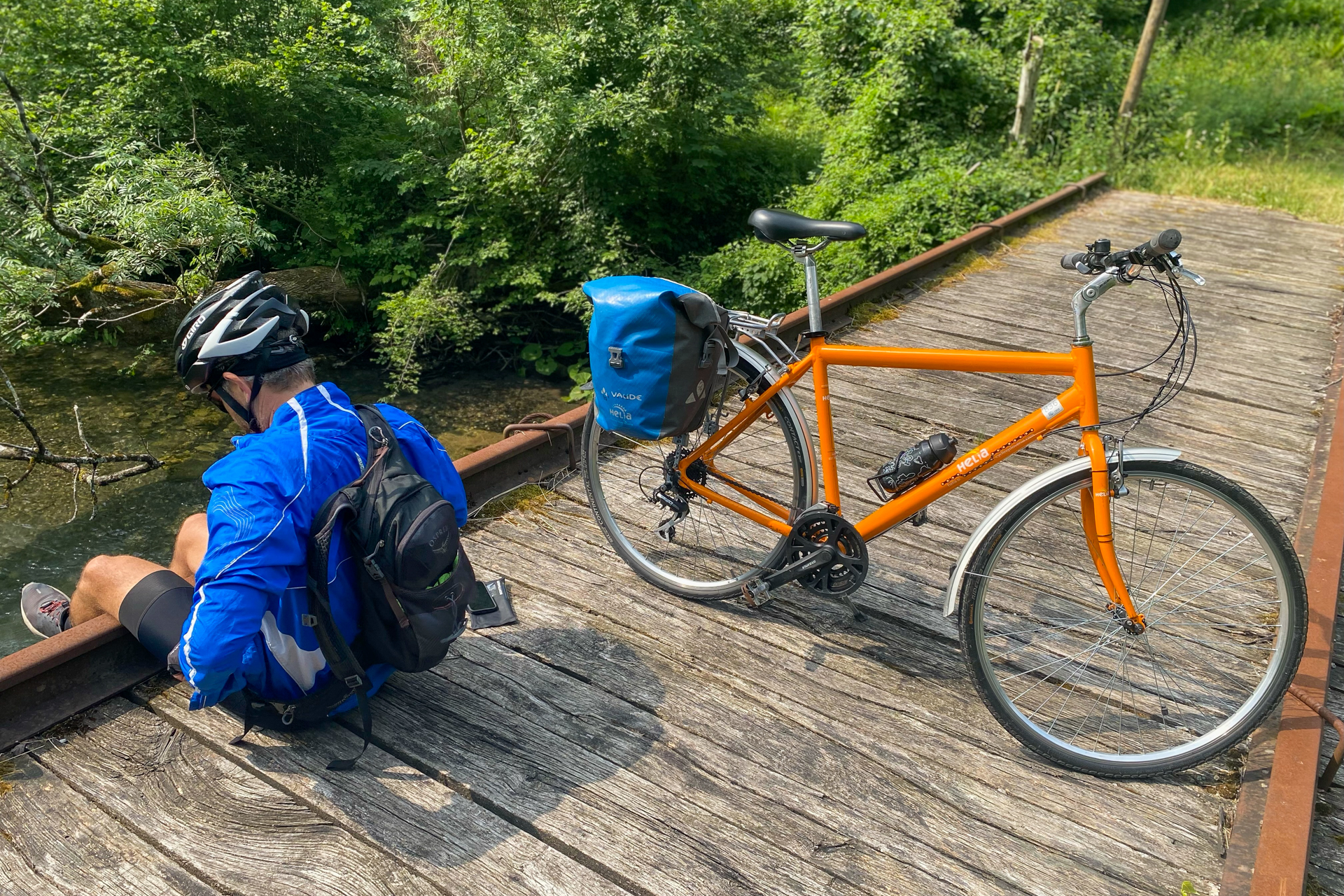 A man sits with his legs daggling over a bridge while an orange bike stands across the bridge.