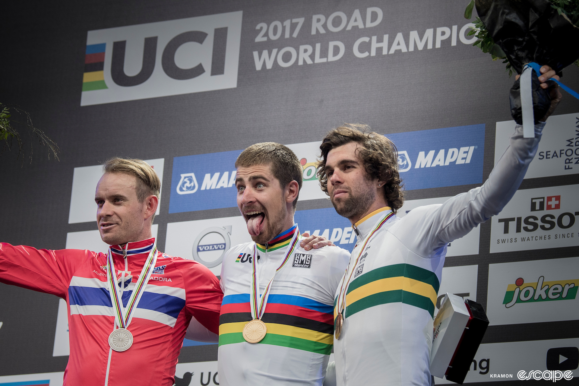 Peter Sagan on the podium in his rainbow jersey, with his tongue out, after winning the 2017 Worlds. He's flanked by Alexander Kristoff and Michael Matthews.