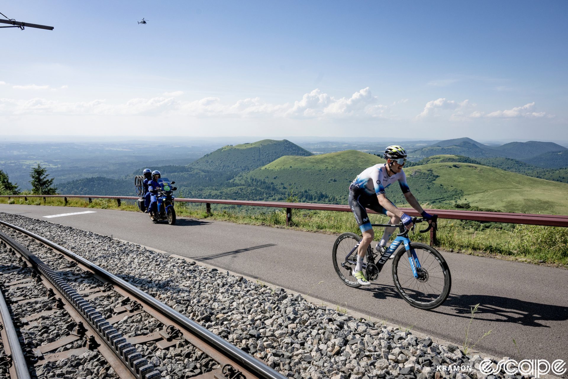 Matteo Jorgenson, all alone on the Puy de Dôme finish climb of stage 9 of the 2023 Tour de France. He's alone in the breakaway, on a road closed to fans. Behind, the valley and low hills of the remnant volcanoes of the Massif Central are seen, green and verdant under a pale blue sky. A lone blue Shimano neutral service moto follows, and the cog summit railway stretches down the road at left.