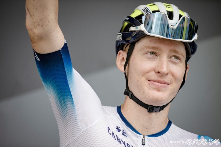 Matteo Jorgenson waves to the crowd at sign-in for the 14th stage of the 2023 Tour de France. He's wearing the special-edition white Movistar team kit, and his sunglasses are tucked into his helmet vent.