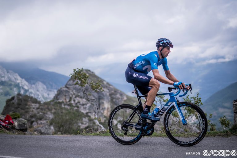 Imanol Erviti climbs in the 2019 Vuelta a España. The Spaniard is alone on the road, a focused expression to match the rugged rocky outcrop behind.