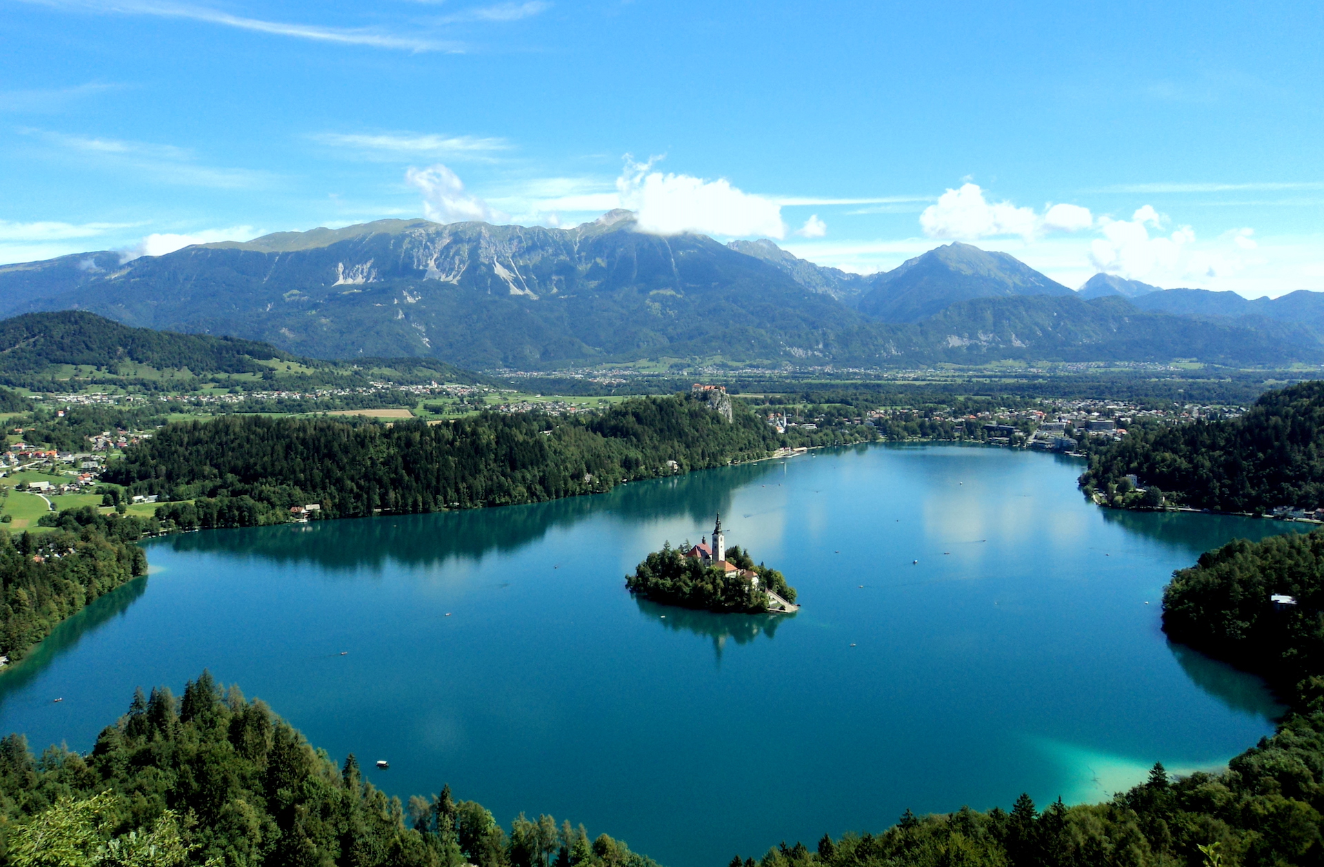 An aerial photo of a stunning, blue lake, with a church-island in the centre and mountains in the background.