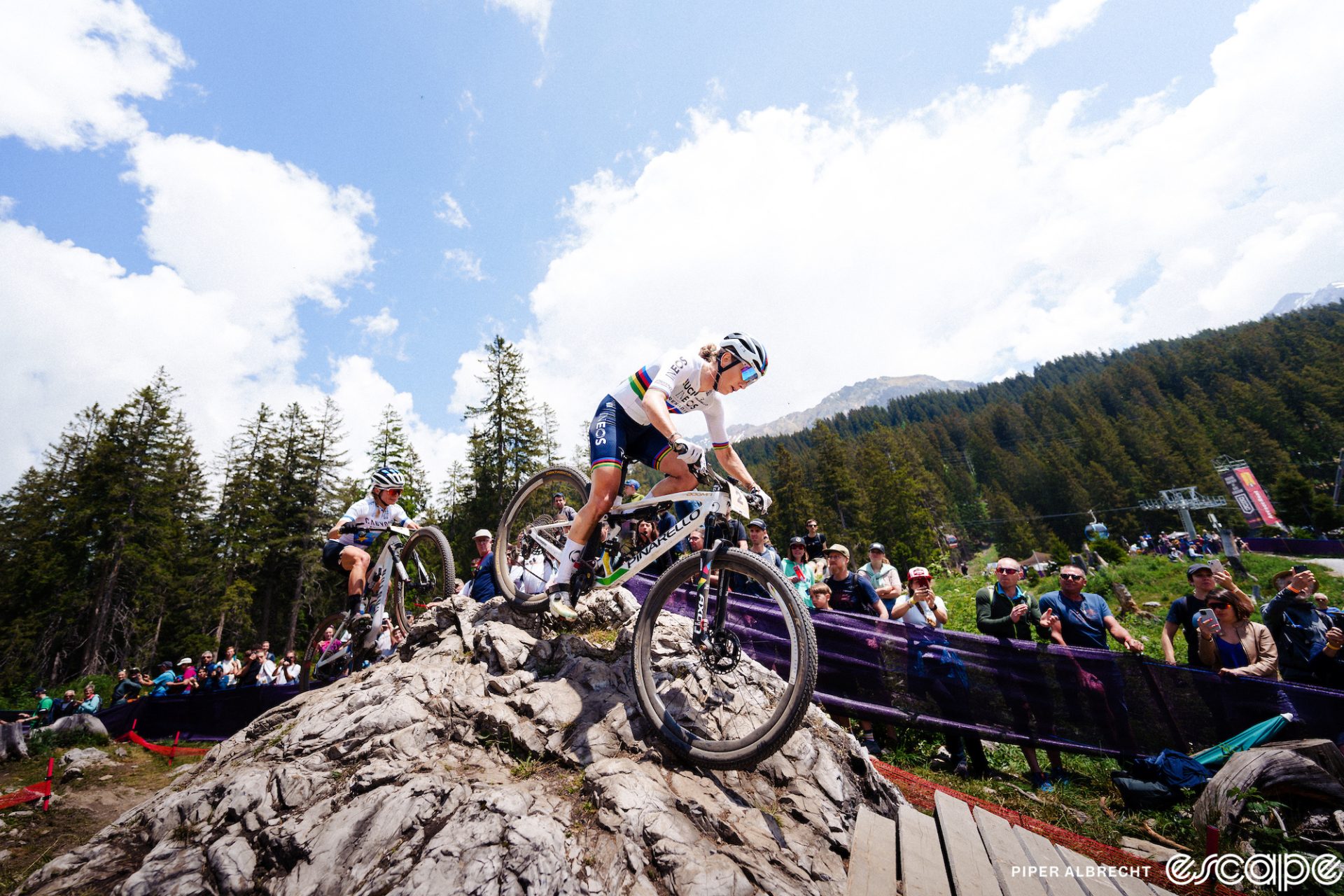 Pauline Ferrand-Prevot rides a rocky section at a World Cup race. She's just cresting the top of the bony outcrop and about to descend onto a wooden bridge. She's wearing the rainbow jersey of World Champion.