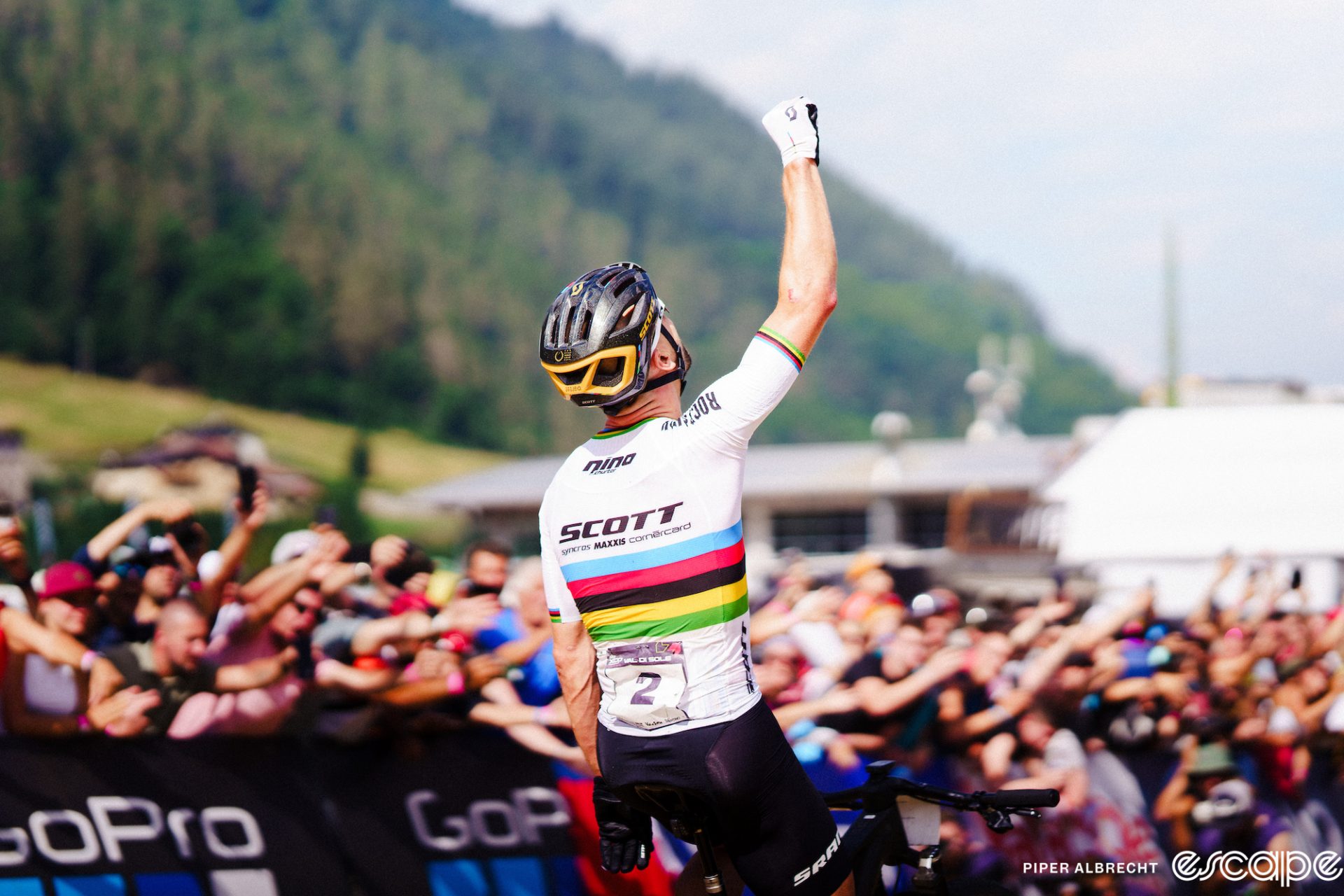 Nino Schurter holds a raised fist to the sky and looks up as he crosses the finish line for yet another World Cup win. He's shown from behind, in the rainbow jersey of World Champion, with big cheering crowds blurred out behind.