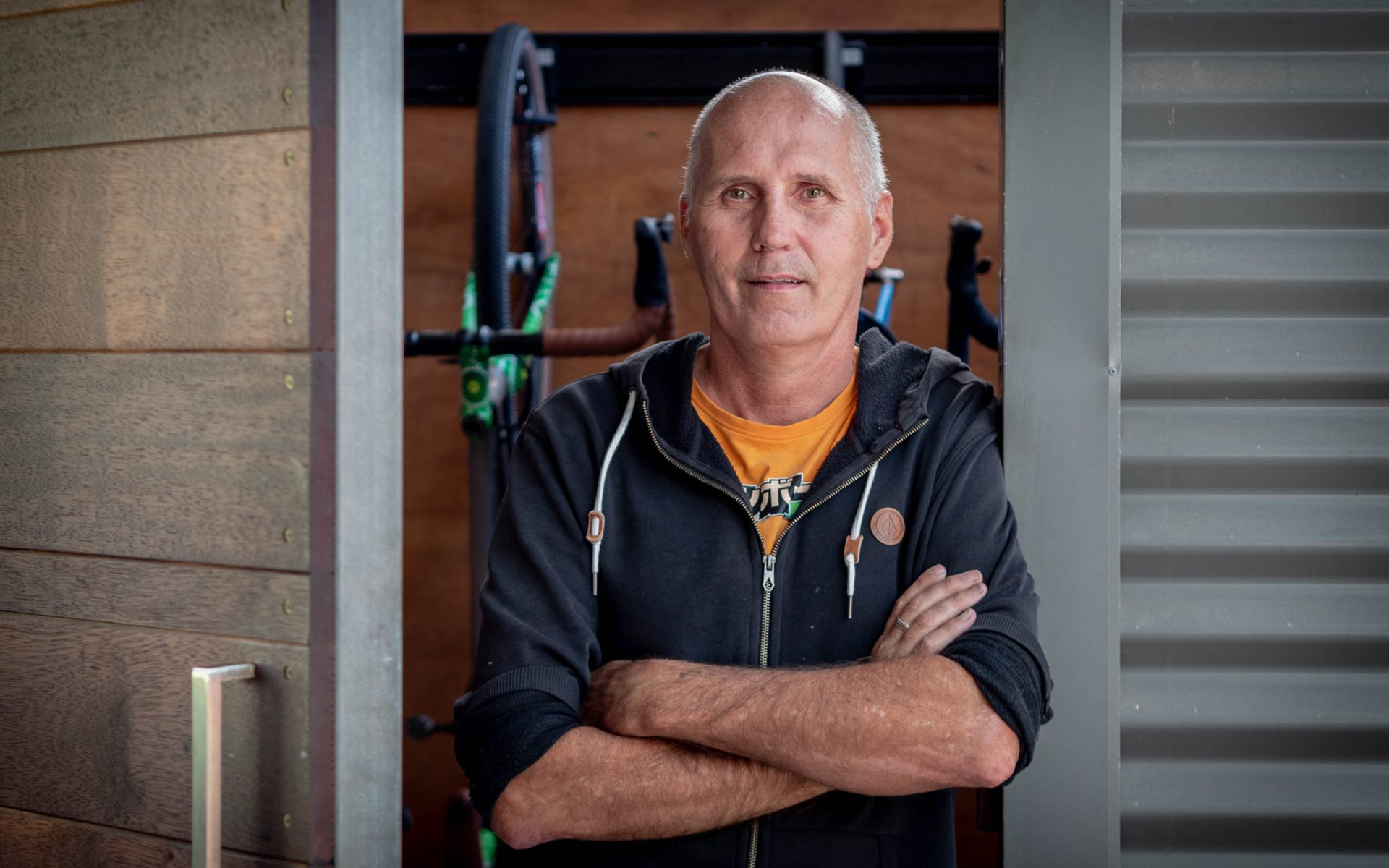 Matt Wikstrom portrait. He stands in a doorway facing the camera, arms crossed, and is wearing a dark grey/black hoodie over a bright yellow t-shirt. In the room behind him are several bikes.