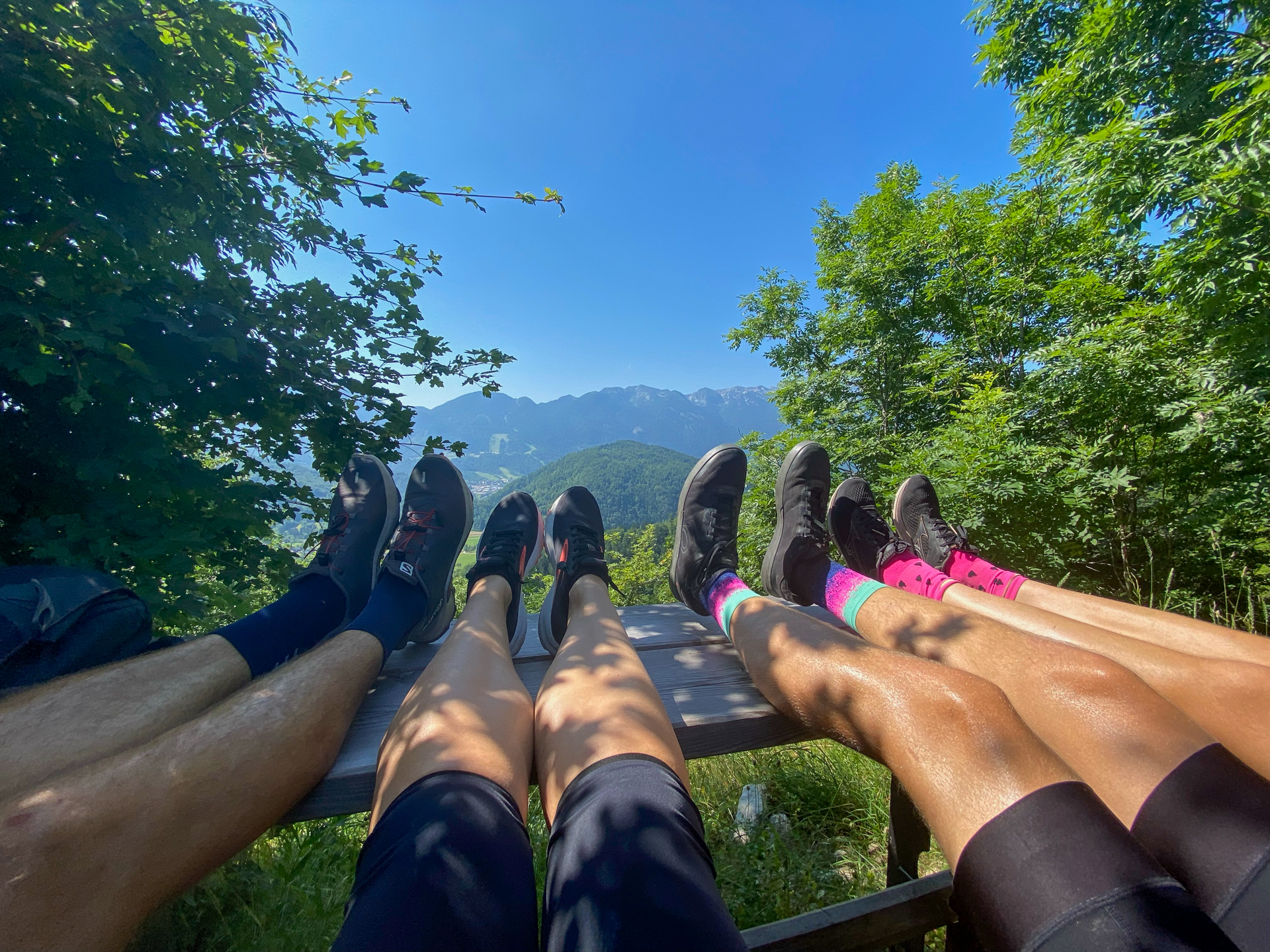 Four sets of sneaker-clad feet rest on a bench with mountains in the background.