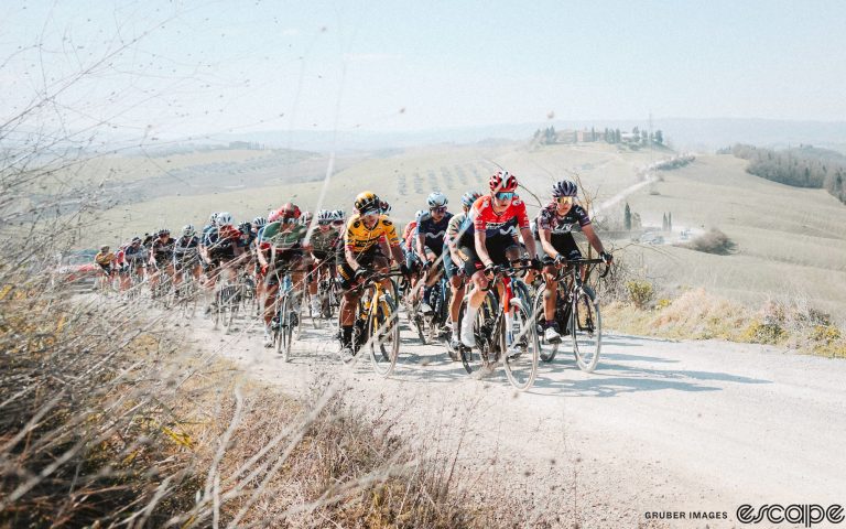 The women's field rides at the 2023 Strade Bianche. The riders are tightly packed, riding a dusty white gravel road with a quintessential Tuscan villa on a hill in the background.