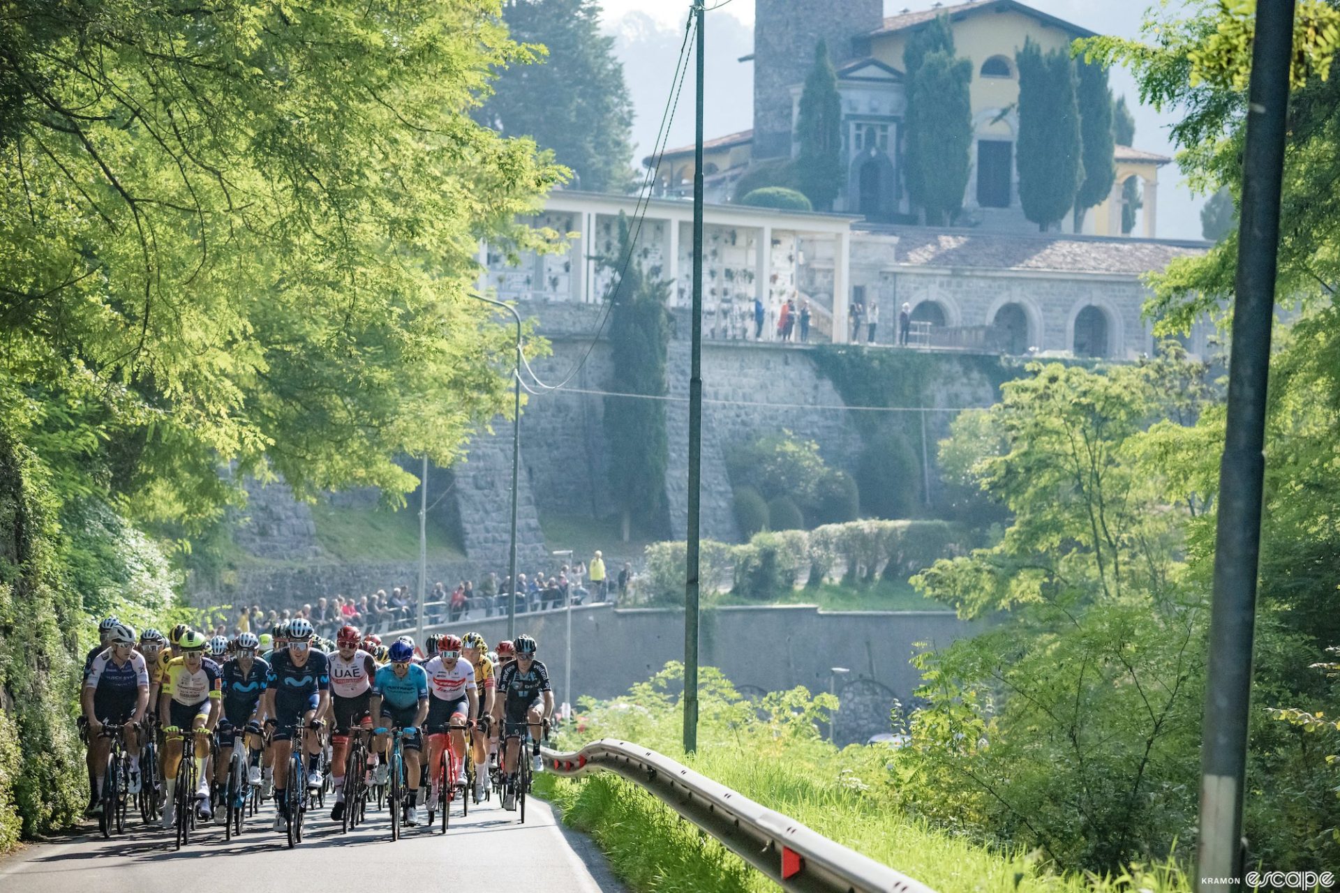 The peloton racing through Lombardy in 2022. The riders are a tight pack, on a road with trees overhanging and a large rock wall and village behind.