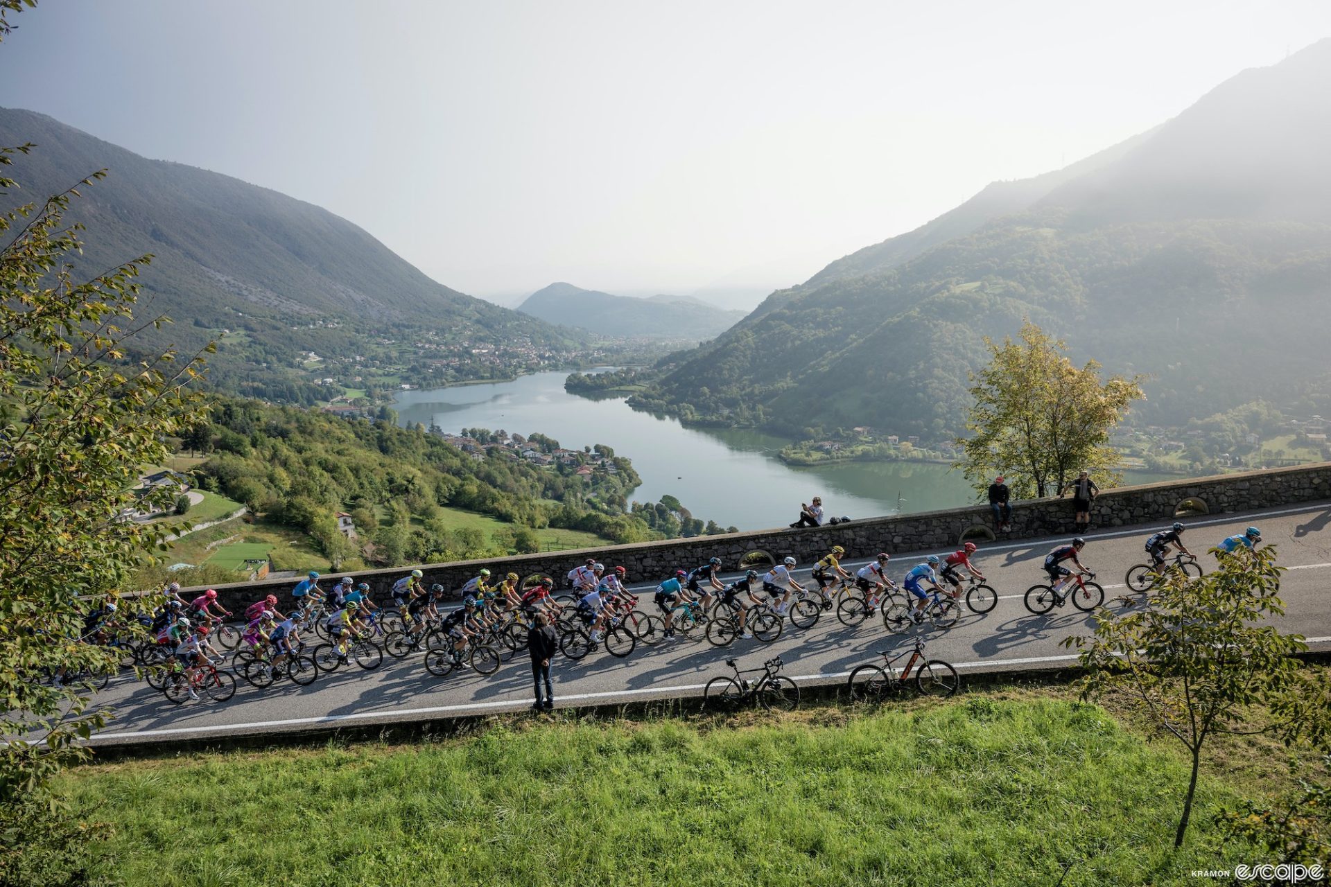 The peloton climbs a road past a lake, with steep, forested hills rising off the shore on either side.