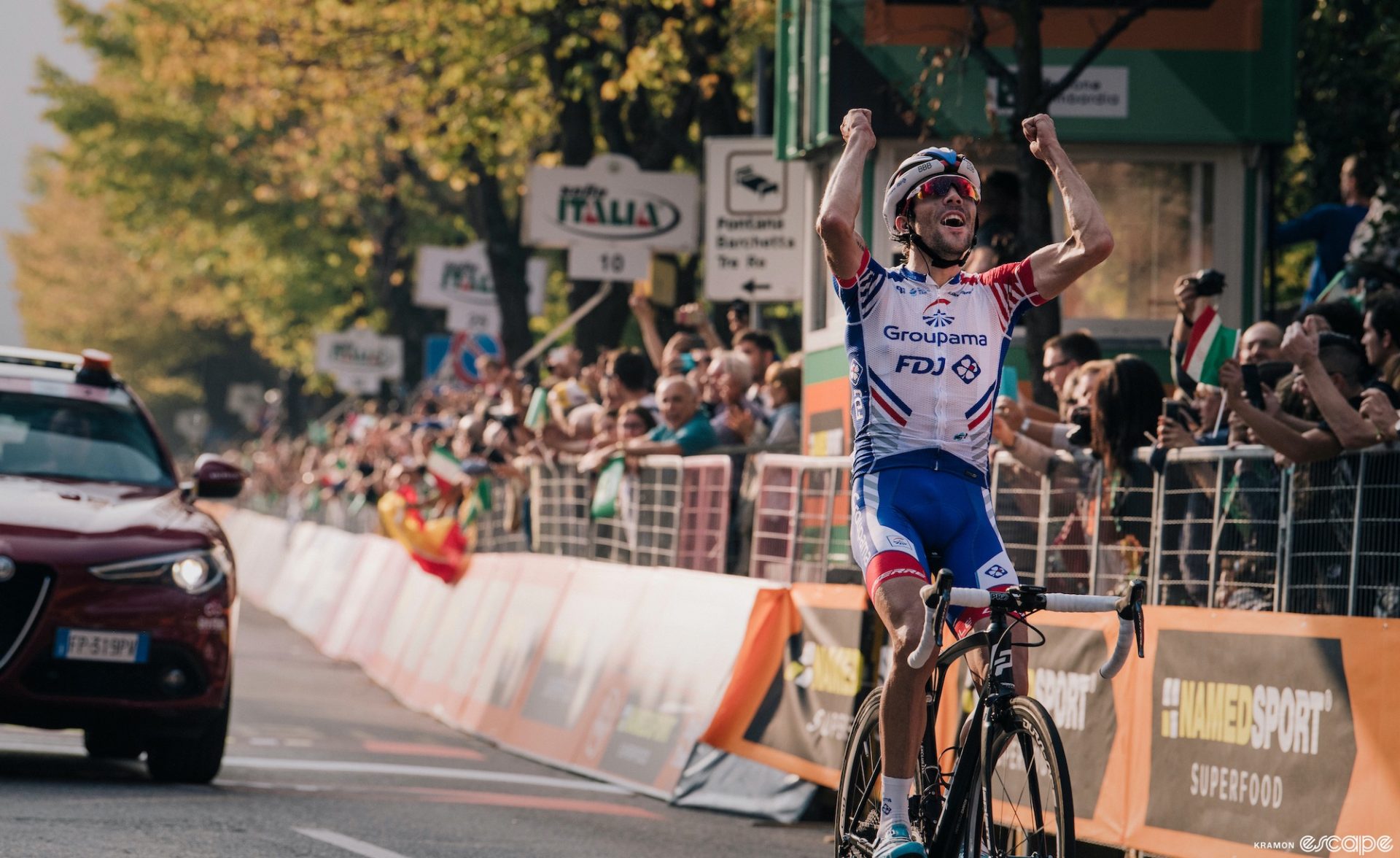 Thibaut Pinot wins Il Lombardia in 2018. The Frenchman is crossing the line alone, arms raised in a closed-fist salute. His head is tilted slightly back and there is an expression of joy on his face.