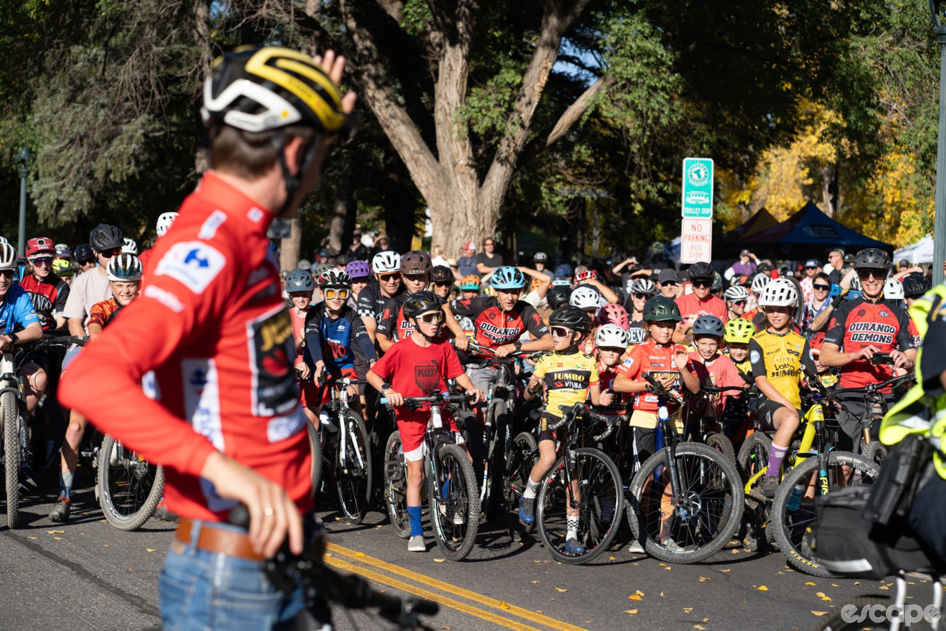 Kuss stands astride a bike, in his red jersey and a helmet, looks back at a crowd of cyclists ready to follow him on a celebratory ride. the front of the massive group is entirely kids, including several dressed in donated Jumbo-Visma  jerseys.