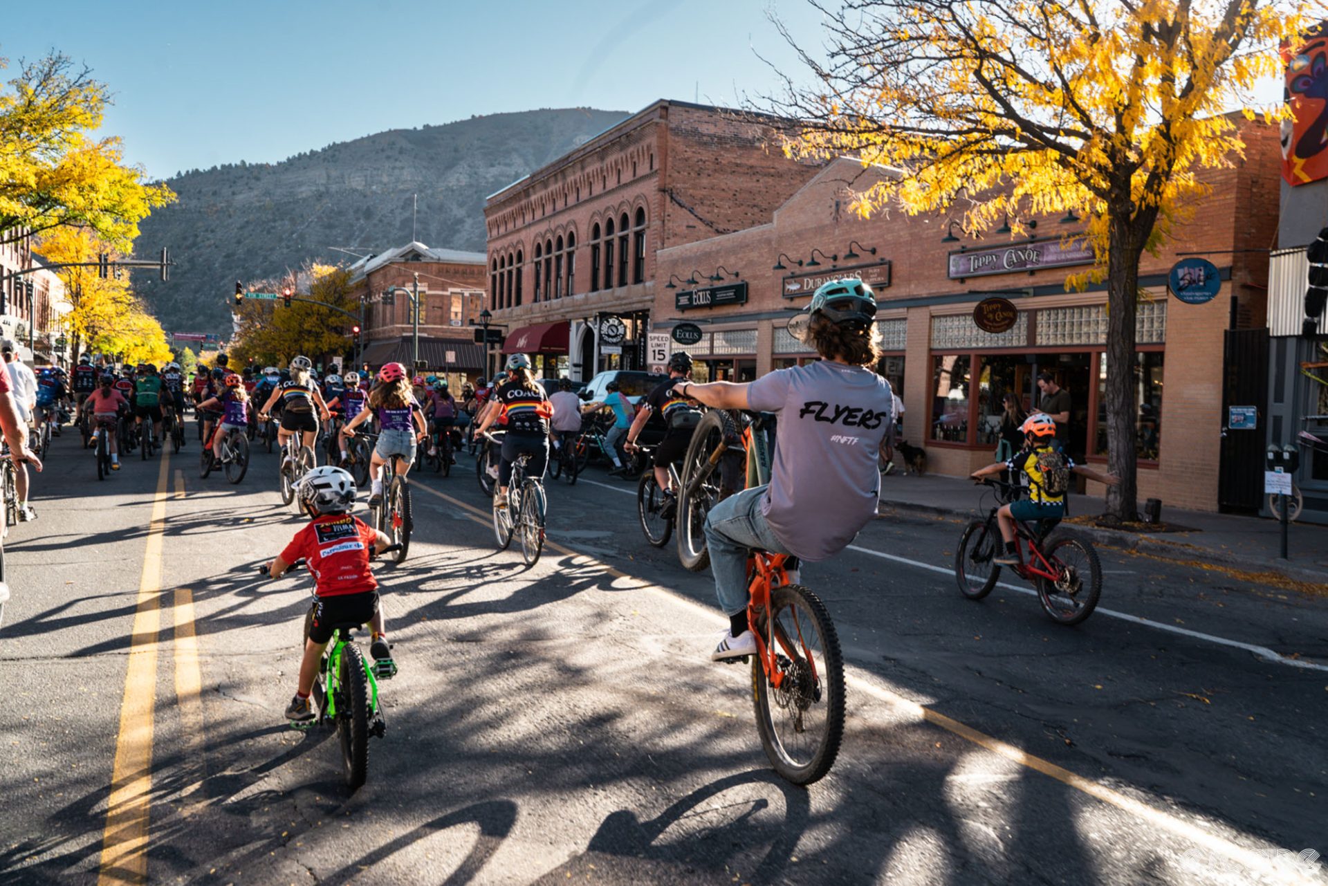 The celebratory ride rolls down a street in Durango. It's gorgeous afternoon sun, yellow leaves on trees, and green forested hills in the background. A rider at the back in a Flyers t-shirt pops a wheelie on his mountain bike as the group rides through town.
