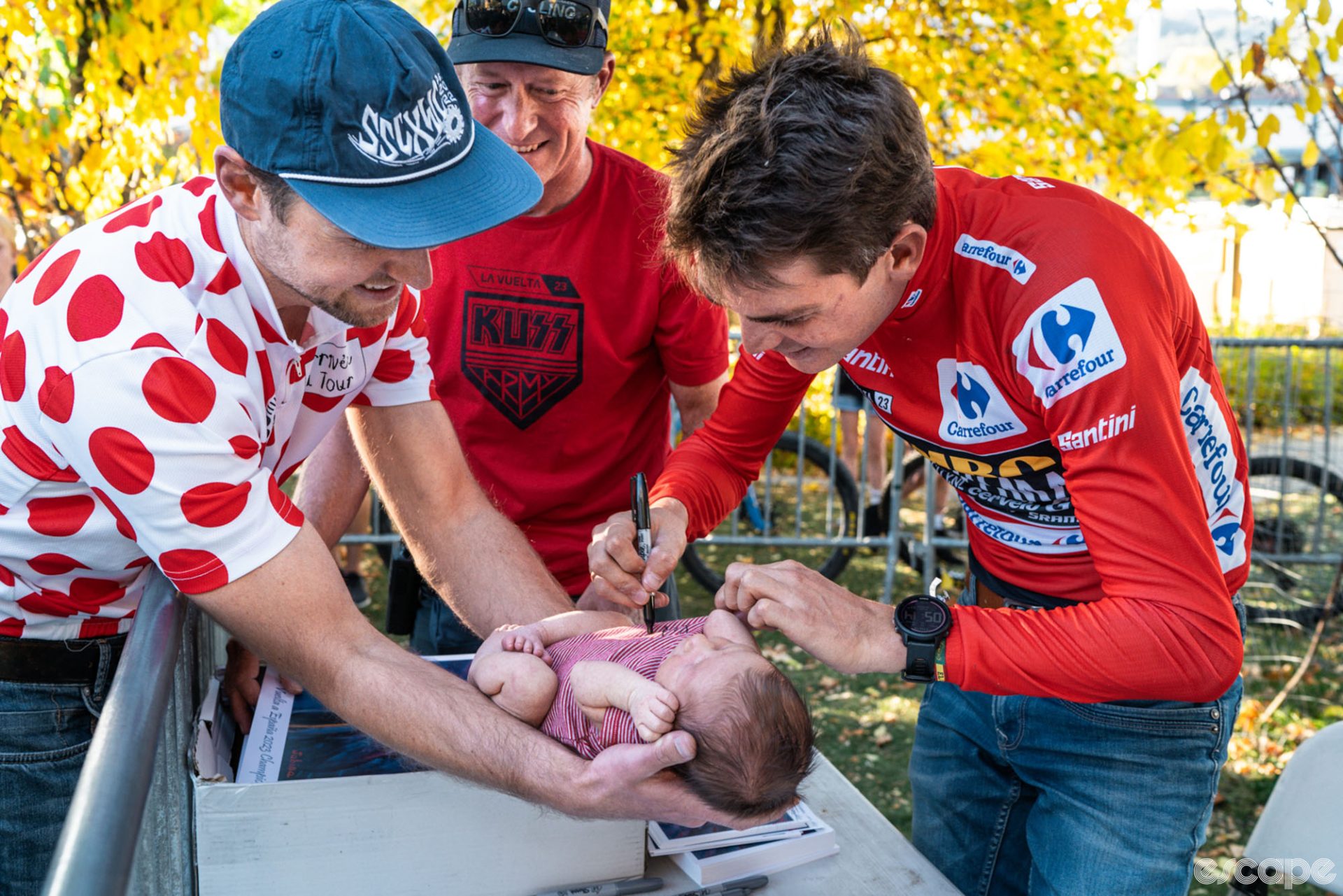 Sepp Kuss smiles as he leans over a table to sign a onesie on a small baby. The child's father is wearing a polka-dot cycling jersey from the Tour de France and holds the child out gently for Sepp's signature.