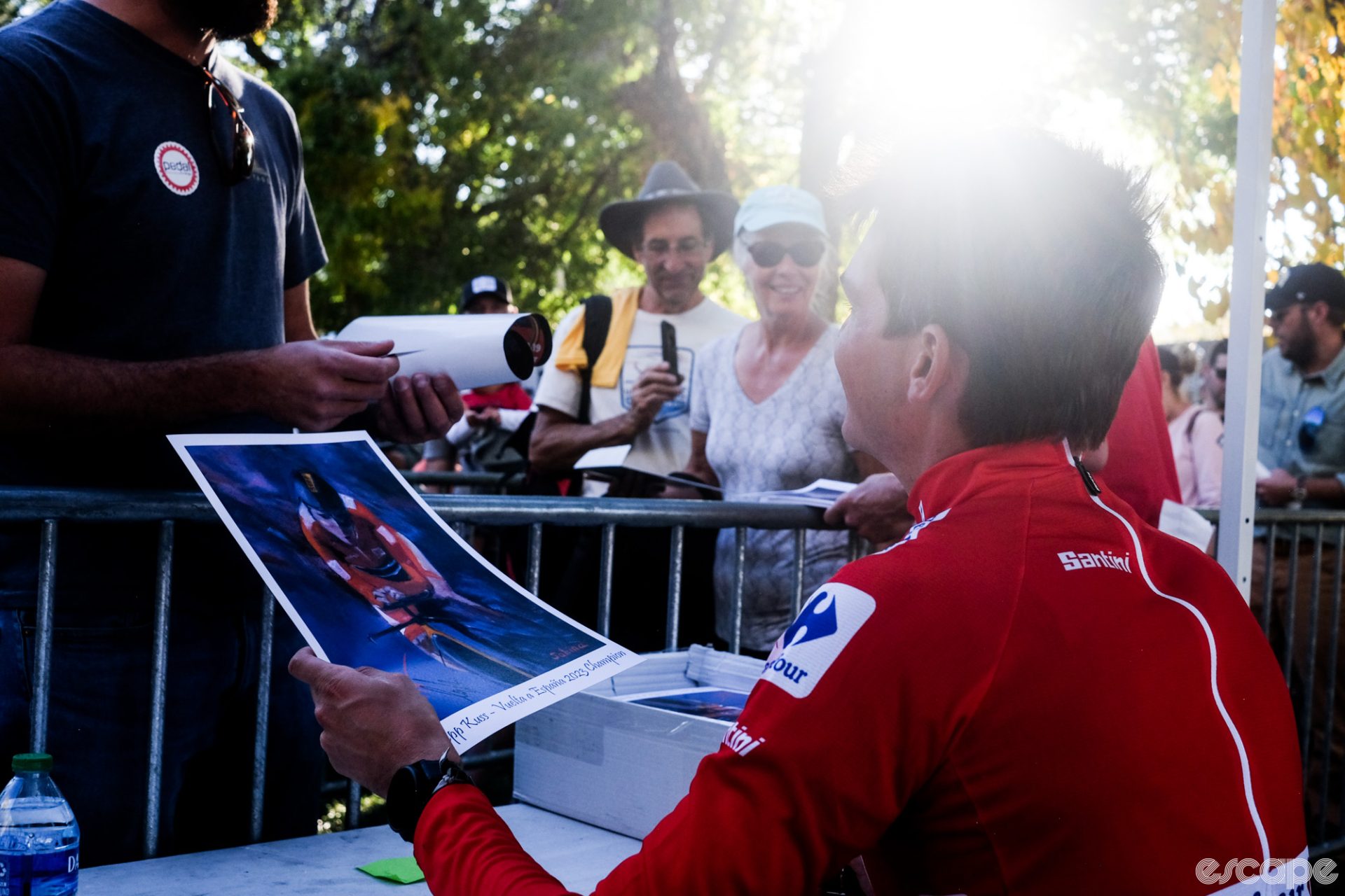 Kuss, in his red jersey, is sitting at a folding table and hands a signed poster to an autograph seeker. More fans wait behind for their turn.