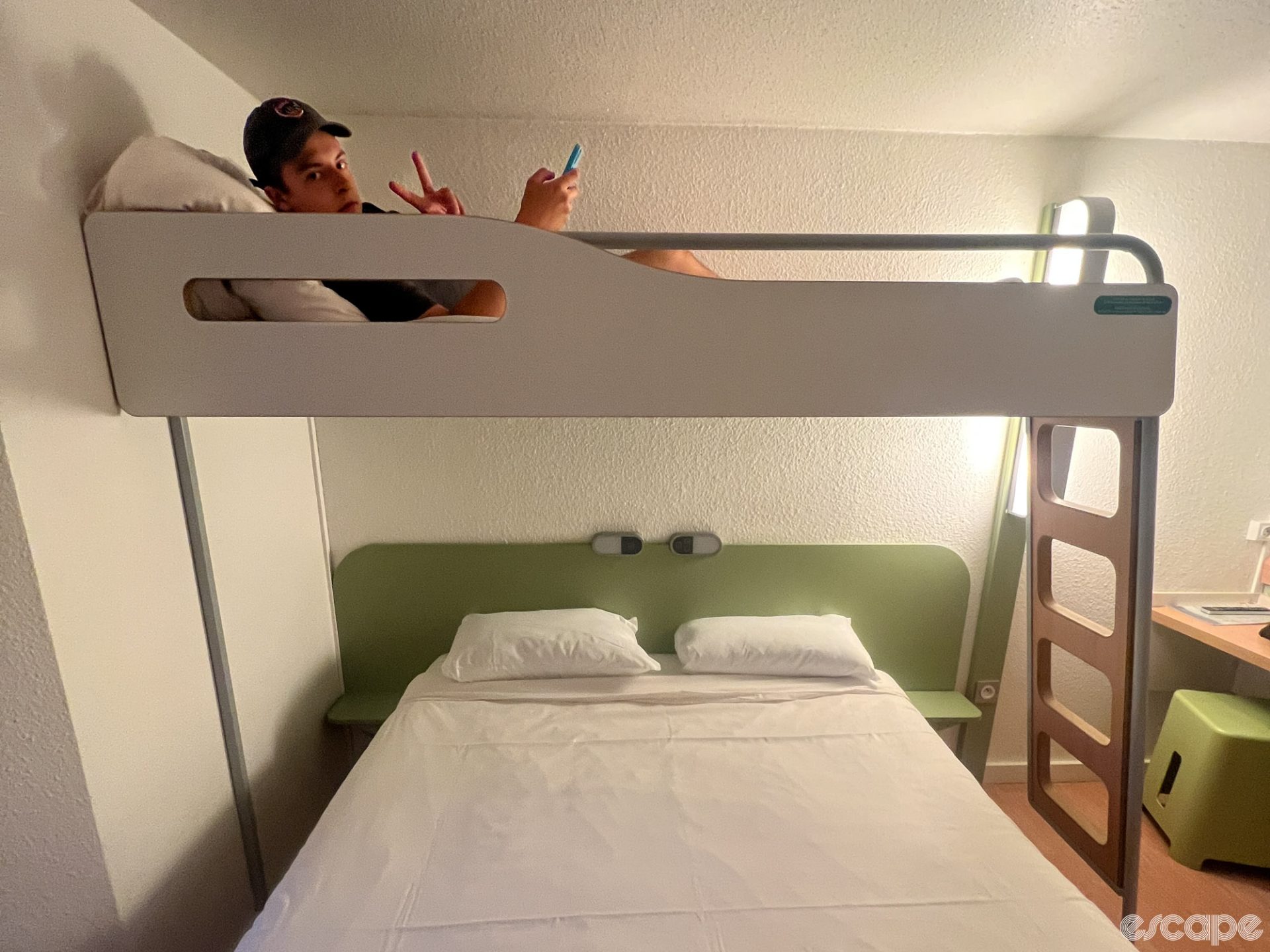 A large man in a small bunk bed gives the V sign. He looks positively thrilled to be staying in a budget hotel.