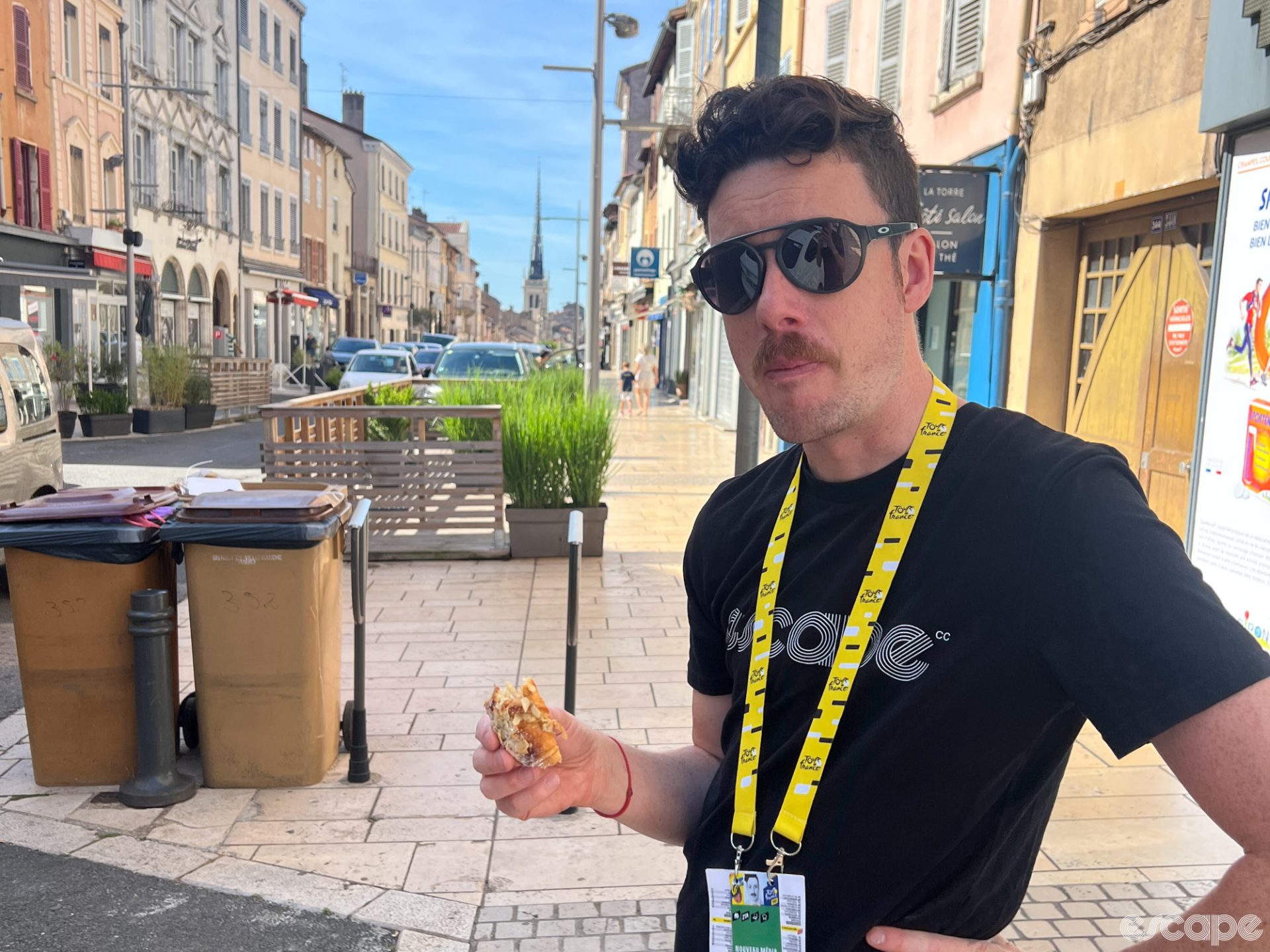 A man who may or may not be Escape's own Iain Treloar enjoys a sandwich on a random street in France. He is wearing very stylish Oakley sunglasses and a black Escape t-shirt and has a bemused expression on his face as if to say, "You're not posting this on the web site, are you?"