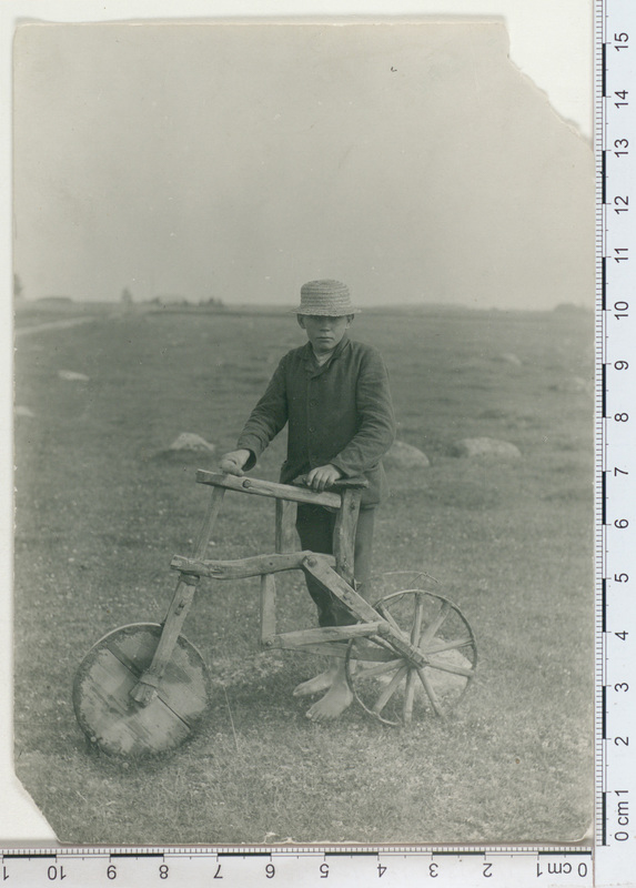 Black and white picture with measurement markings along the sides. Two corners are torn. It shows a boy in a hat wearing a suit, with an rustic-looking wooden bike. 