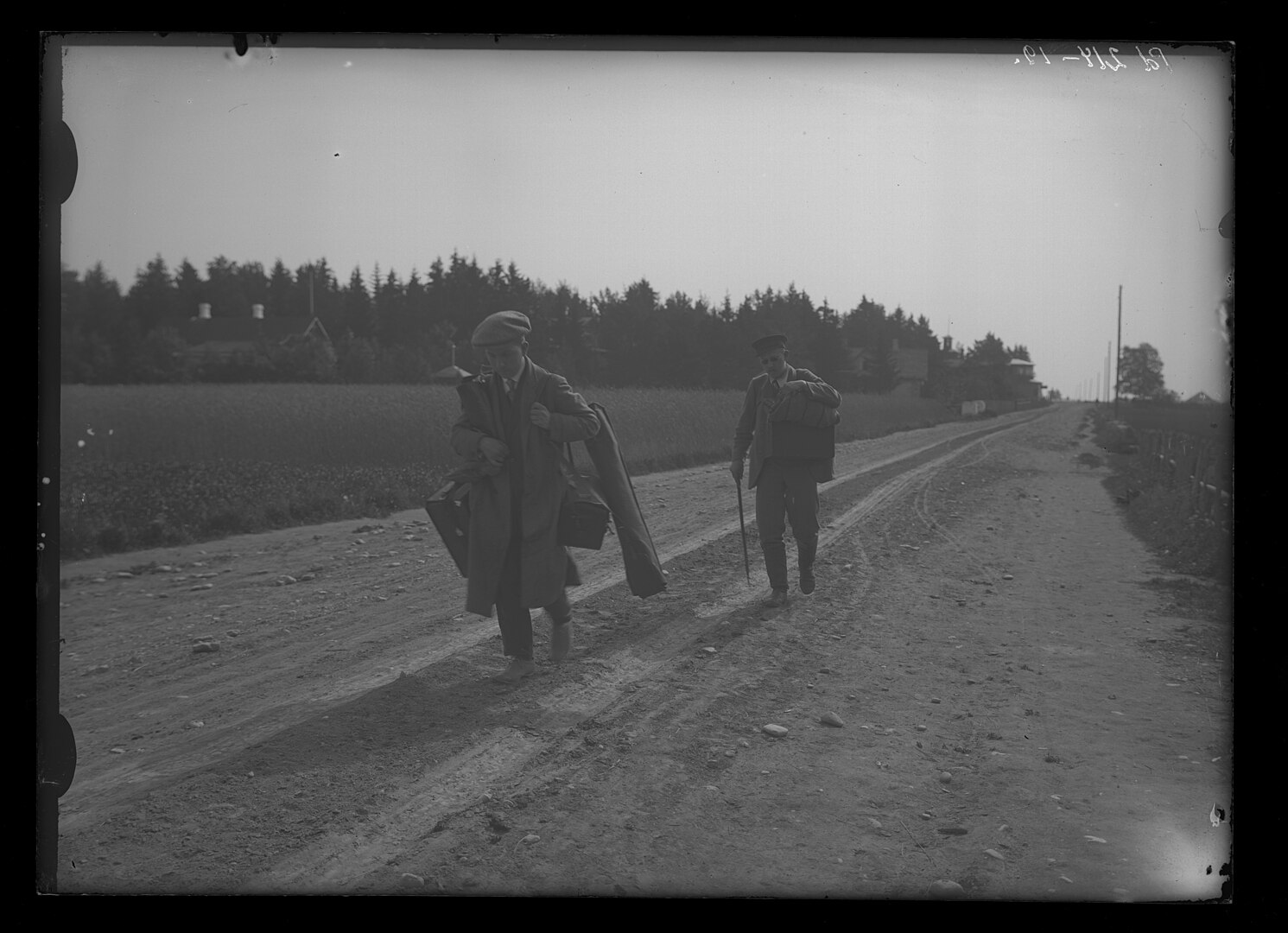 Black and white picture: Pääsuke and his assistant trudge along a dirt road in rural Estonia. They are both heavily laden with bags and equipment.