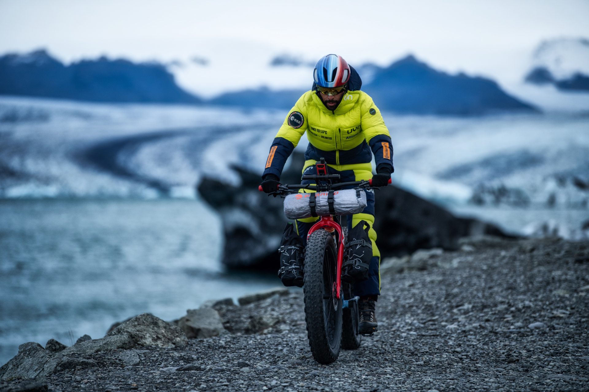 Omar Di Felice rides across a rugged, snowy landscape. He's on bare rock, but there's ice and snow all around and he's dressed warmly in a yellow down jacket. The bike is loaded with supplies.