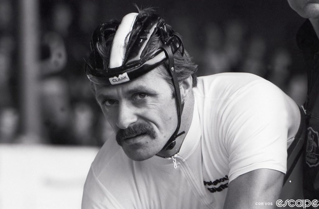 Danny Clark profile. He's looking at the camera with an intense stare, wearing a hairnet-style helmet. His signature bushy mustache completes the tough-guy look.