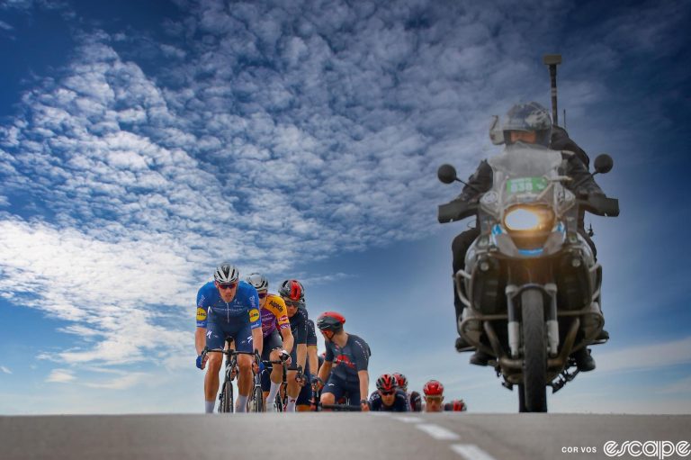 A TV camera moto leads a pack of riders over a rise at the 2021 Tour de France. The moto and riders are softly lit against a blue sky flecked with white clouds.