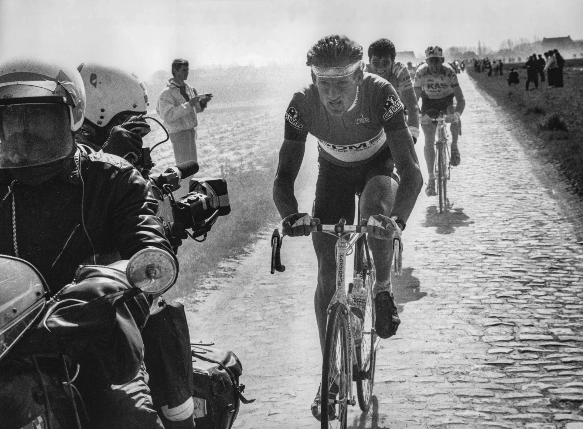 Another old edition of Paris-Roubaix, where Adrie van der Poel (wearing a large, 80s-era headband and no helmet) leads a line of cyclists on dry, dusty cobbles.