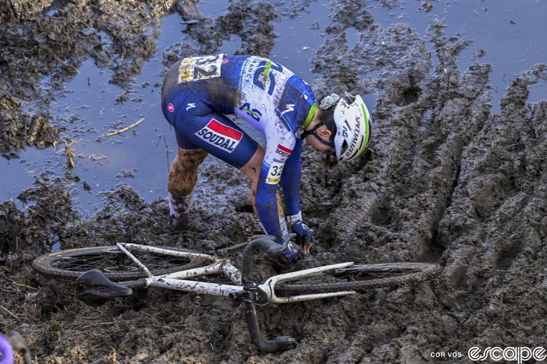 Margot Marasco leans over to try to fix her right shoe at the Superprestige Niel. She's standing in deep, watery mud and her shoe is covered in muck.
