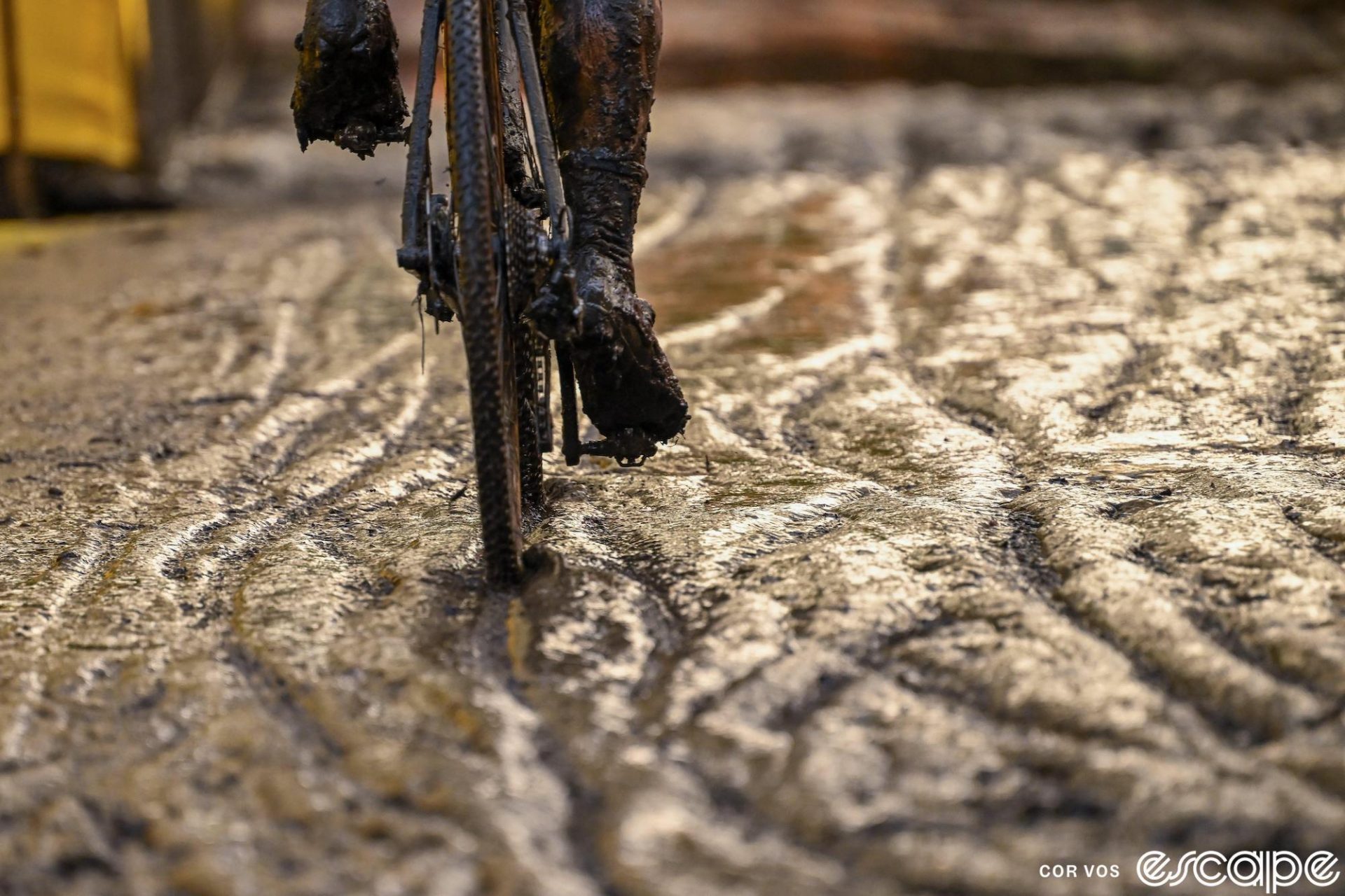 From behind, a rider is seen riding through mud at the Superprestige round at Niel. Only the wheels and feet are visible, covered in mud as the bike traces another track.